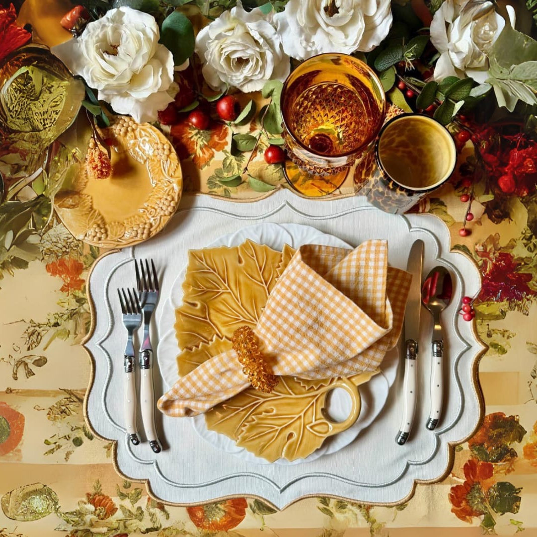 A set of embroidered fabric placemats in a cream color, presented on a dining table setting with plates, cutlery, and a floral tablecloth.