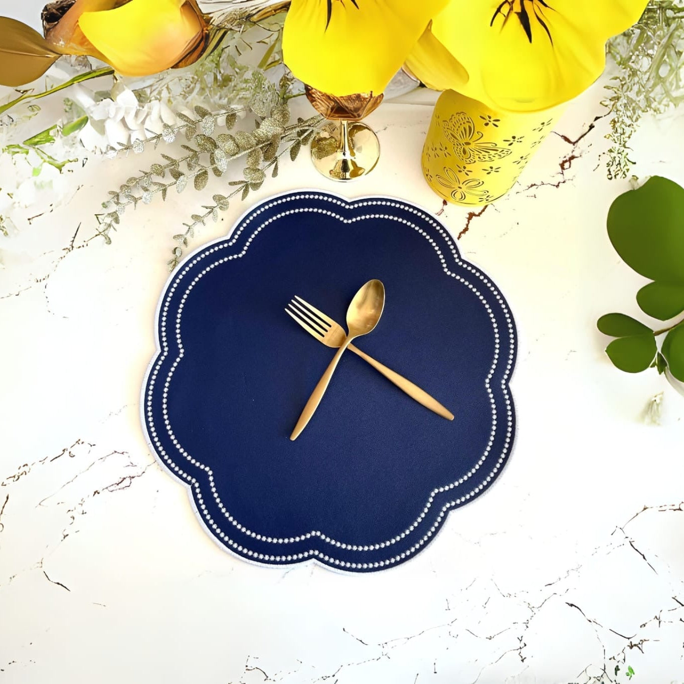 A round faux leather placemat in blue with white embroidery, displayed on a table with a fork and spoon on top, surrounded by yellow flowers and green foliage.