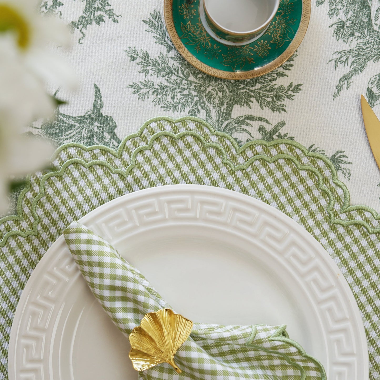 A set of green and white checkered placemats and napkins displayed on a dining table, with gold cutlery and a floral patterned tablecloth in the background.