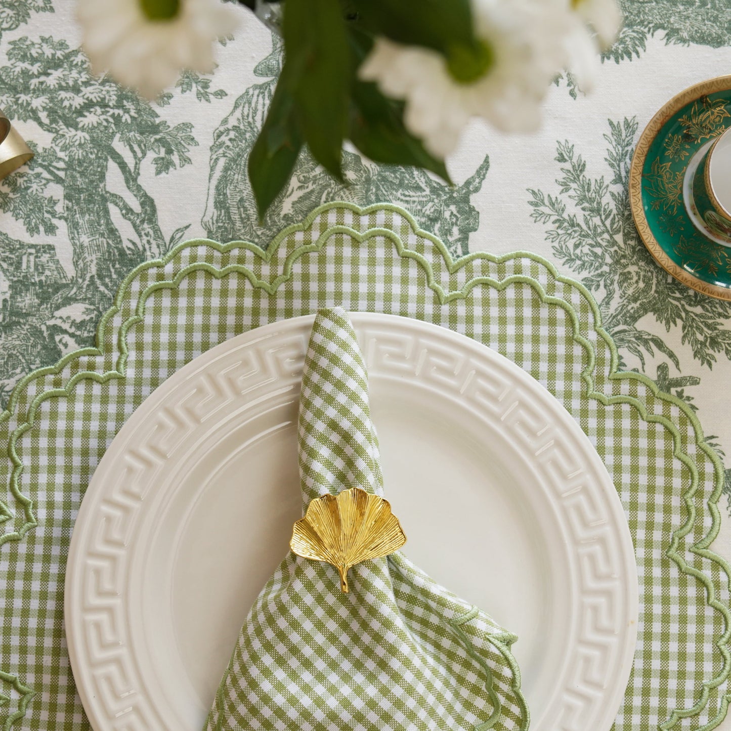 Dining table setting with green toile tablecloth, white plates, and decorative napkins.