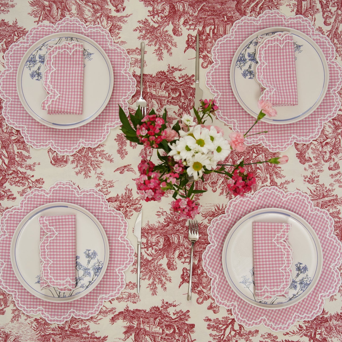 A set of pink and white gingham placemats with floral embroidery displayed on a table with a floral patterned tablecloth.