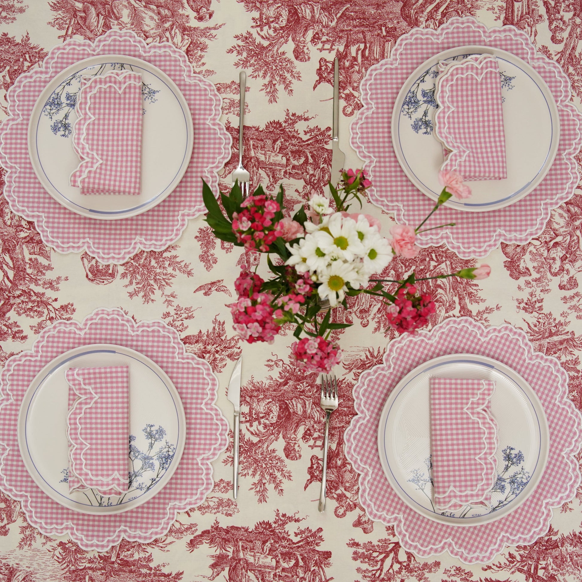 A set of pink and white gingham placemats with floral embroidery displayed on a table with a floral patterned tablecloth.