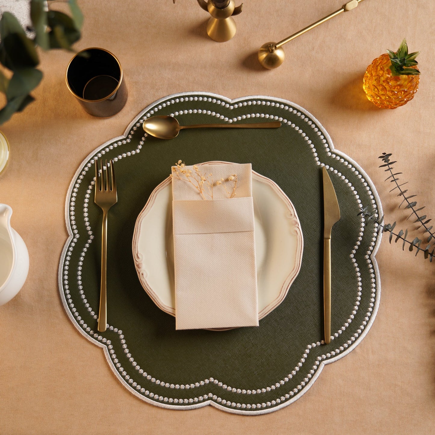 A green faux leather placemat with a decorative edge, featuring white embroidered details, is displayed on a table setting with a plate, cutlery, and a white tablecloth.