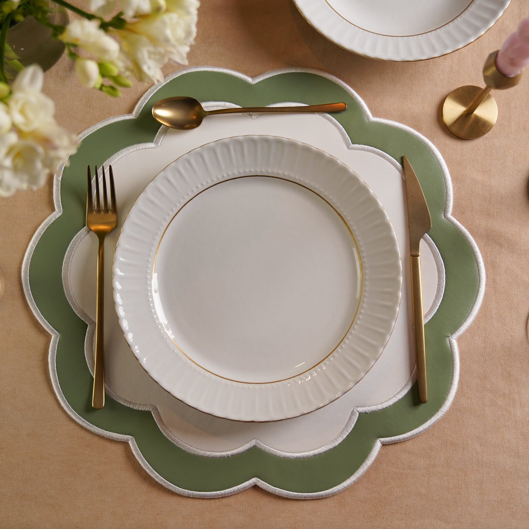 A set dining table with a white and green faux leather placemat, a white plate, and silverware. The table setting is accompanied by glasses and a floral arrangement in the background.