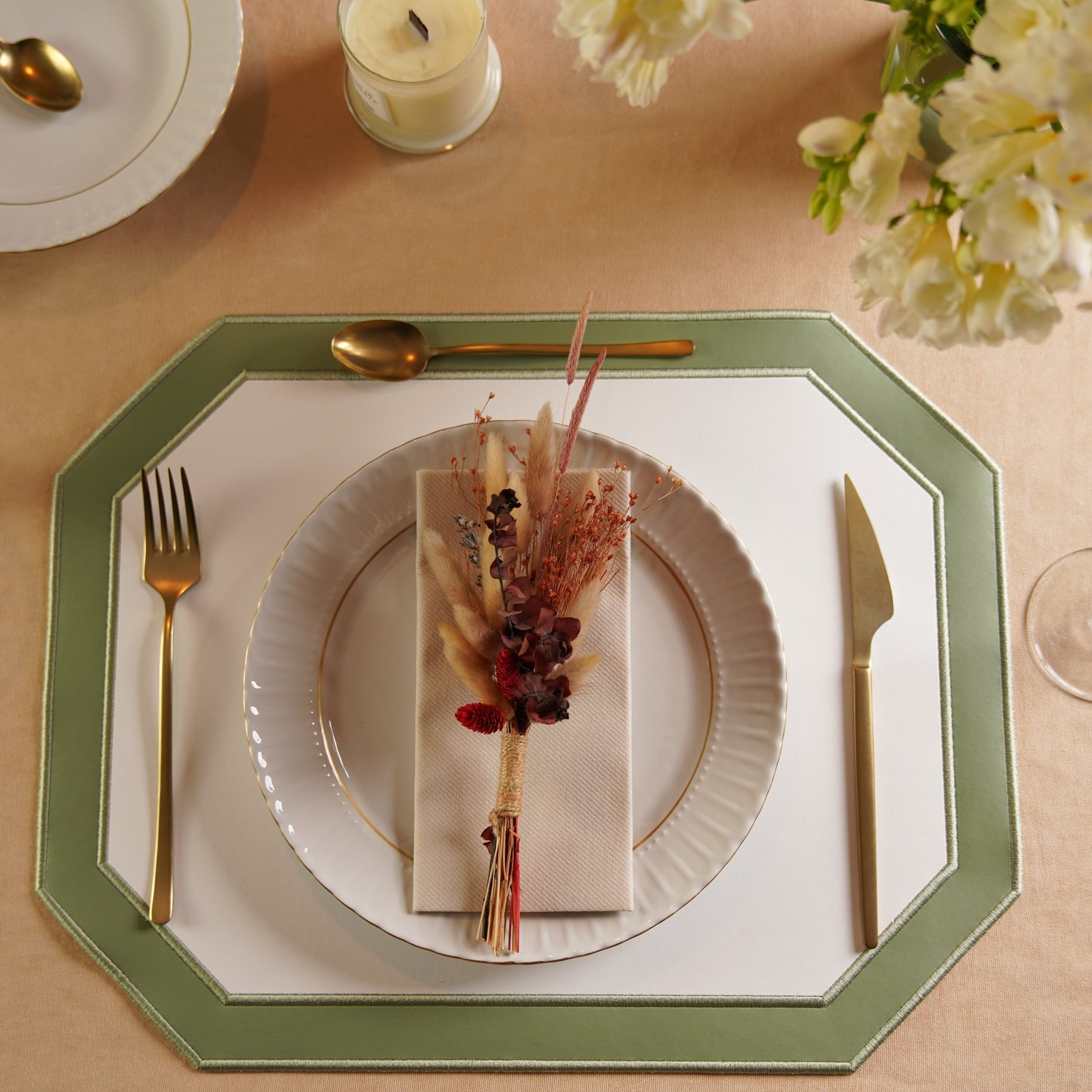 A set dining table with a white and green faux leather placemat, surrounded by plates, cutlery, and a glass. The table setting is complemented by a decorative floral arrangement in the background.