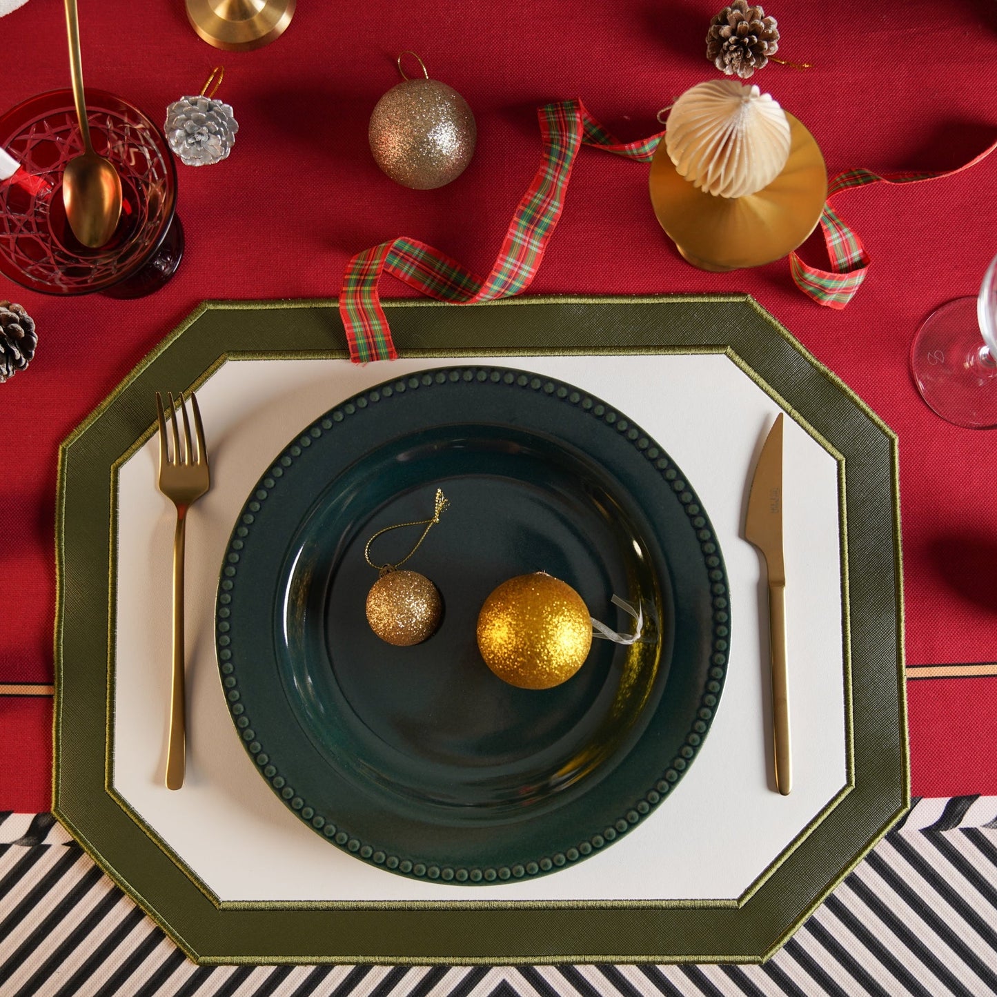 A festive table setting with a dark green faux leather placemat, white plate, and gold and red decorative elements.
