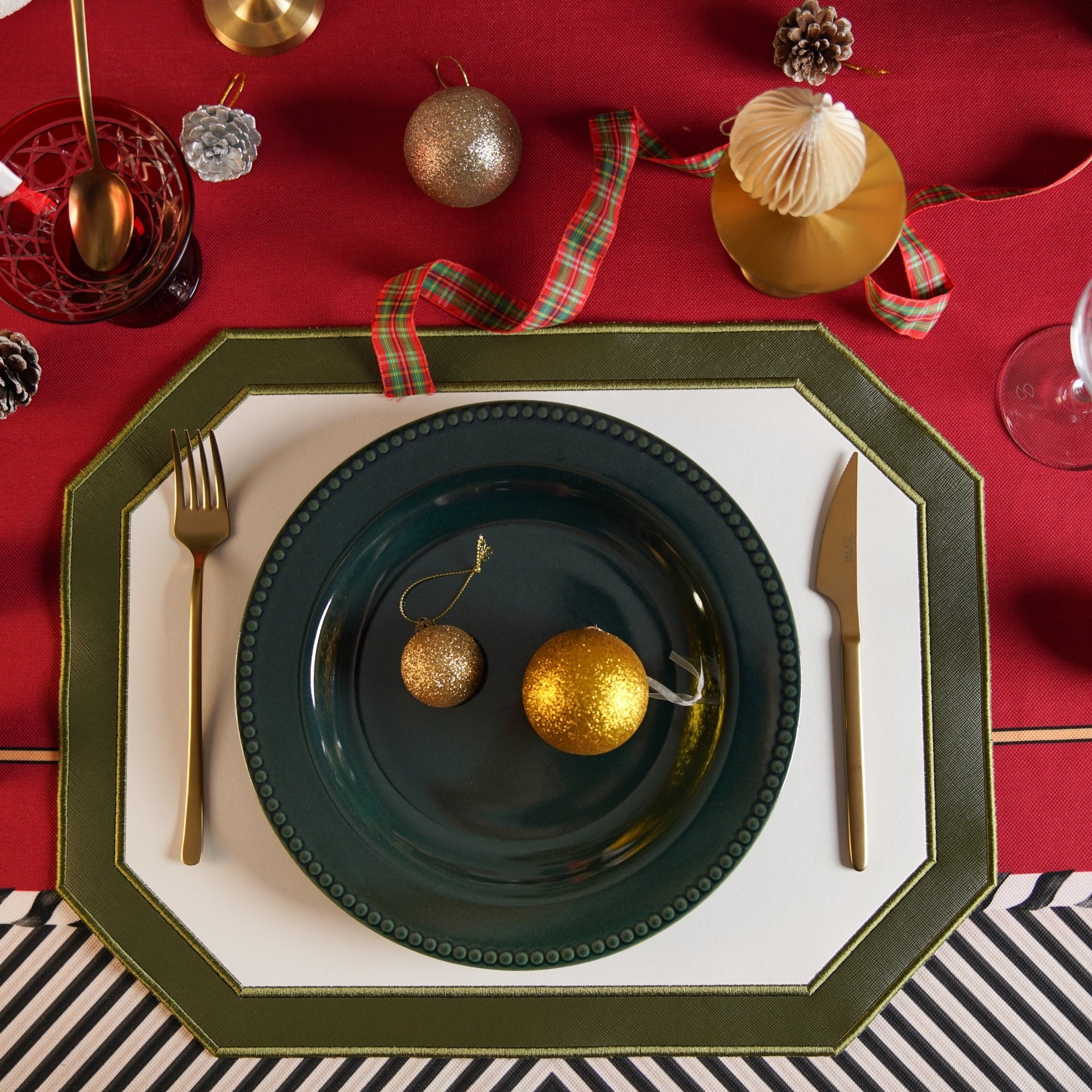 A festive table setting with a dark green faux leather placemat, white plate, and gold and red decorative elements.