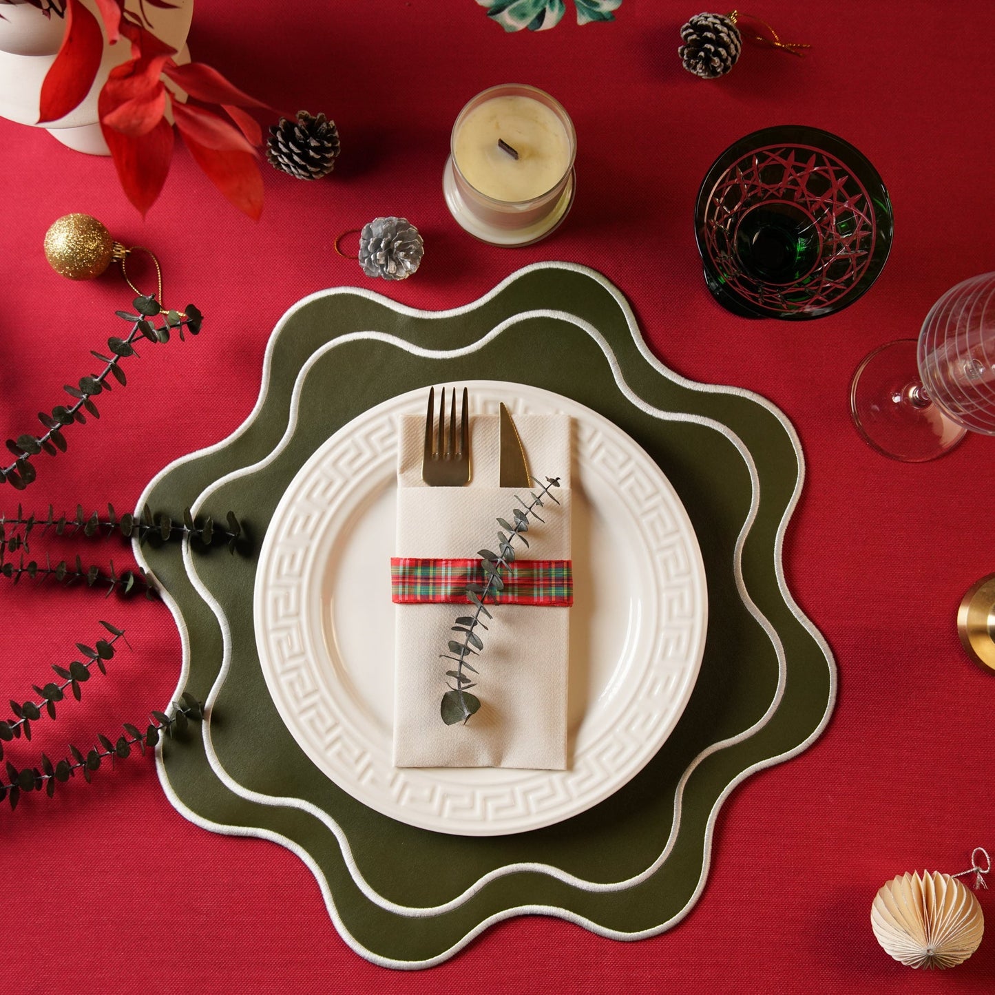 A dark green faux leather placemat with a scalloped edge design, displayed on a red tablecloth. There is a white plate with a fork and knife setting, and decorative elements including candles, a red bow, and green foliage.