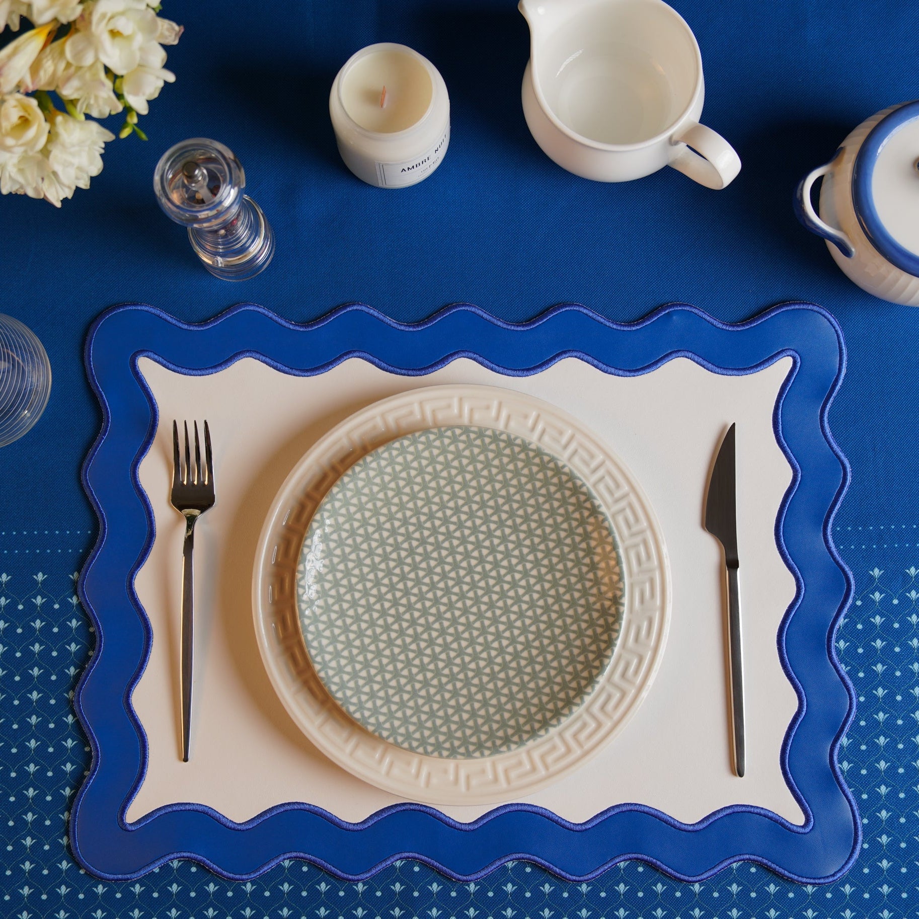 A blue faux leather placemat with white embroidered details, displayed on a table with a plate, cutlery, and a floral patterned tablecloth.