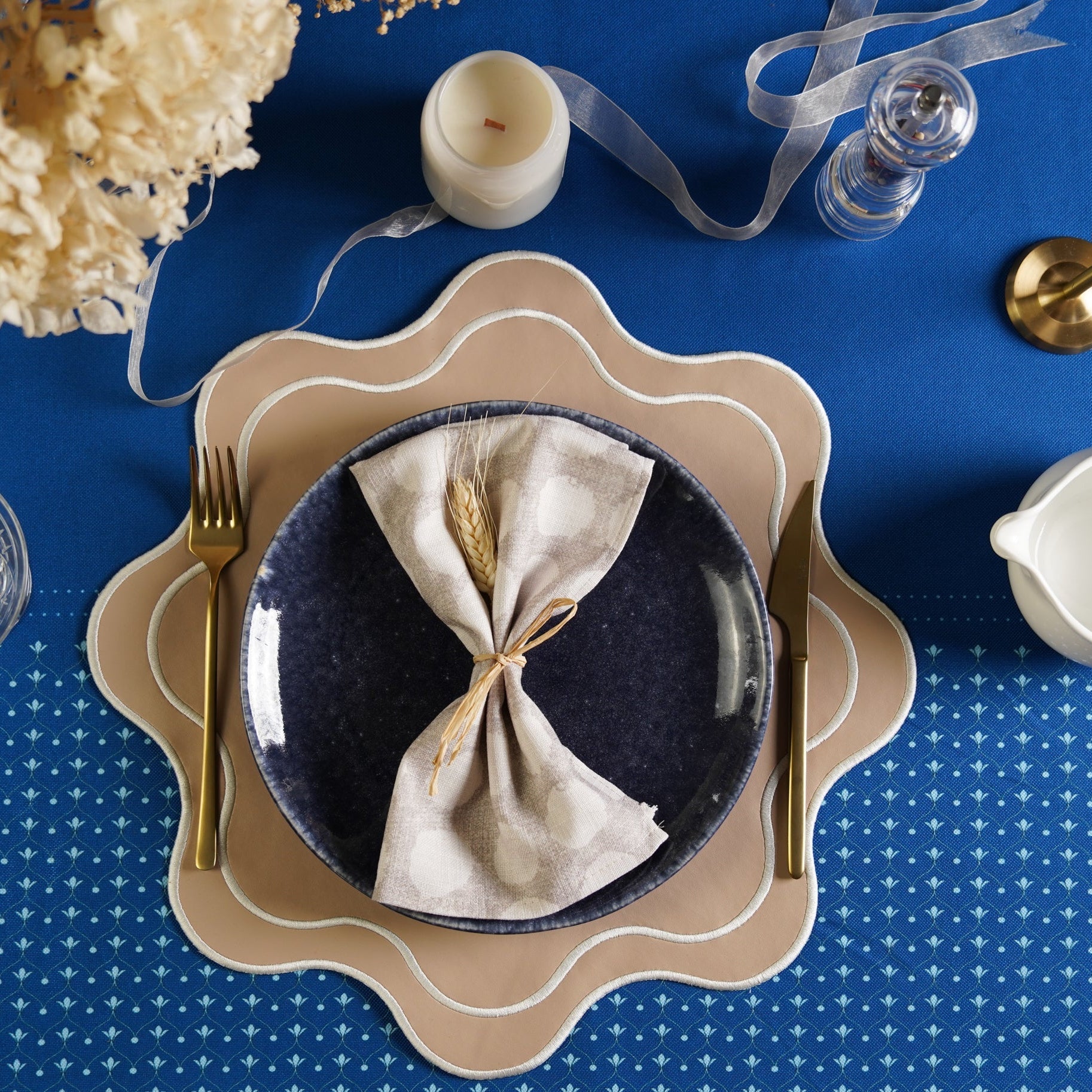 A beige faux leather placemat is laid on a dining table setting, surrounded by a blue tablecloth, a plate, cutlery, and a glass. Decorative elements such as a candle and a vase are also visible in the background.