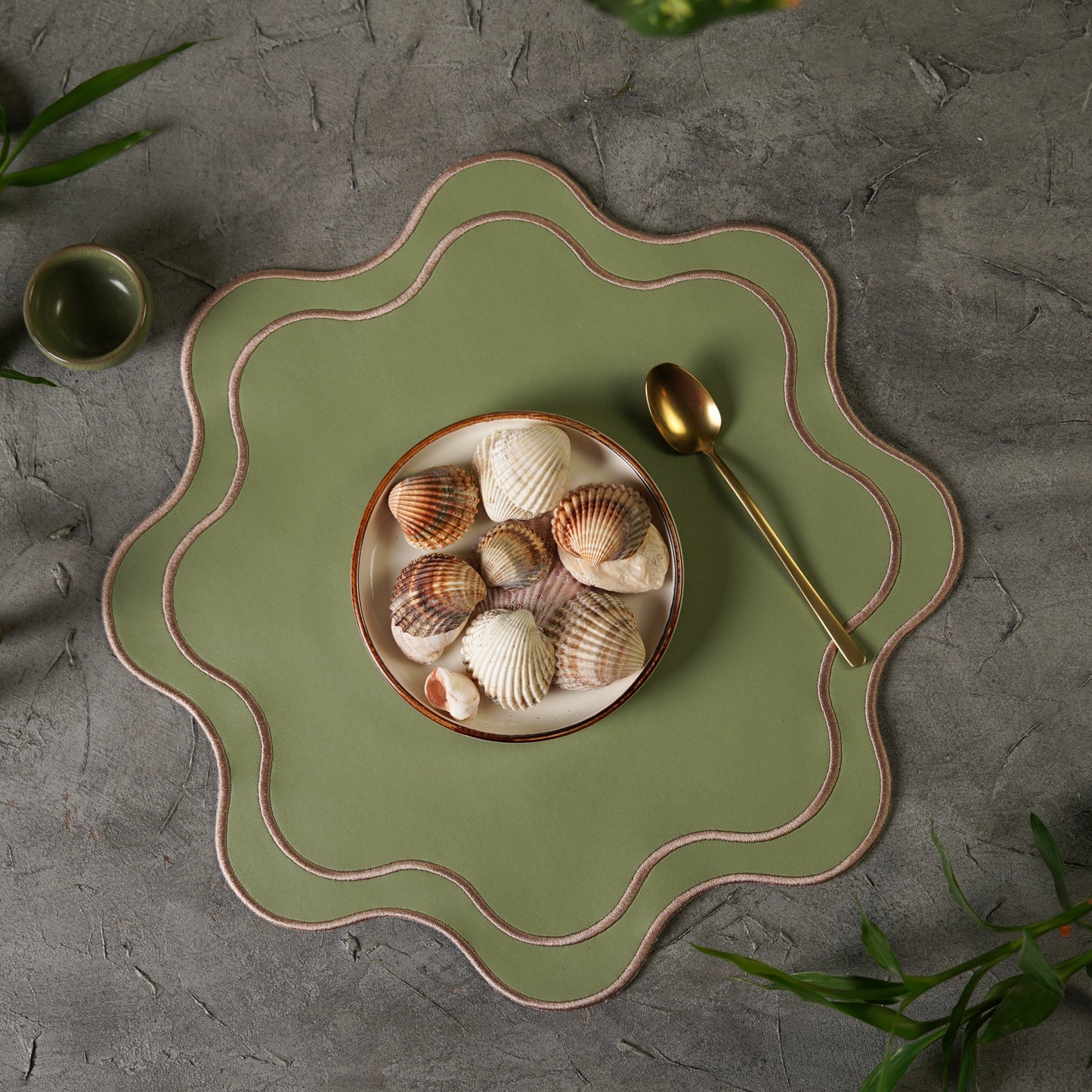 A light green faux leather placemat with a decorative edge, displayed on a table with a bowl and spoon set on top, surrounded by green foliage.