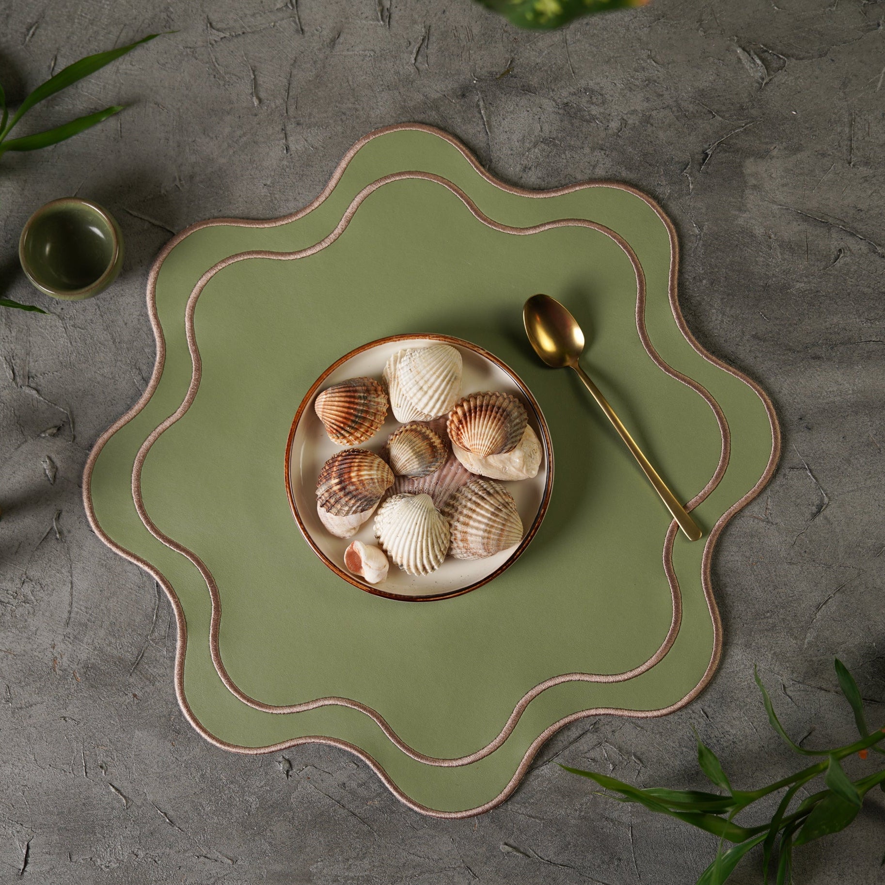 A light green faux leather placemat with a decorative edge, displayed on a table with a bowl and spoon set on top, surrounded by green foliage.