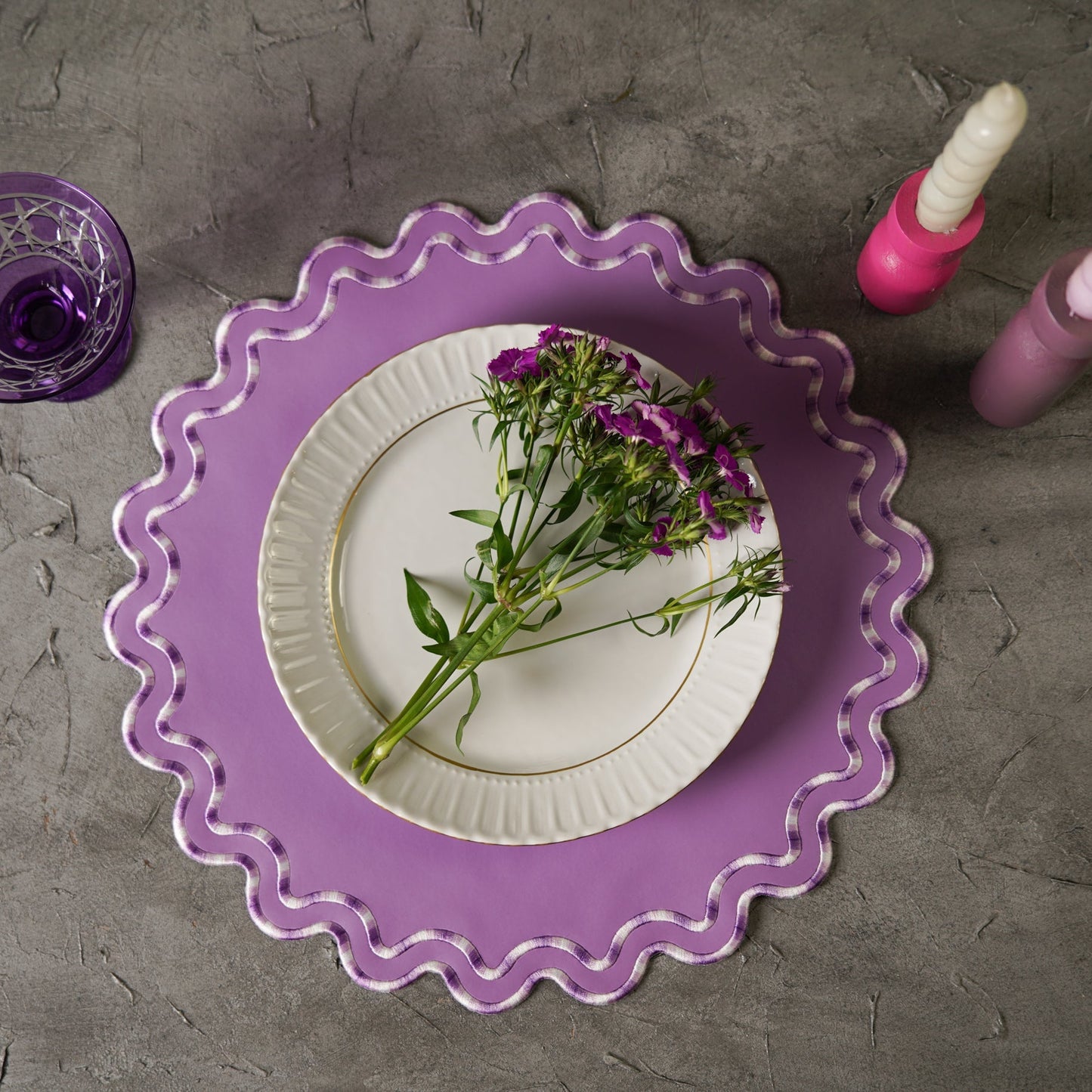 A purple faux leather placemat is laid out on a table, with a white plate on top of it featuring a purple flower. There are also pink candle holders visible in the image.
