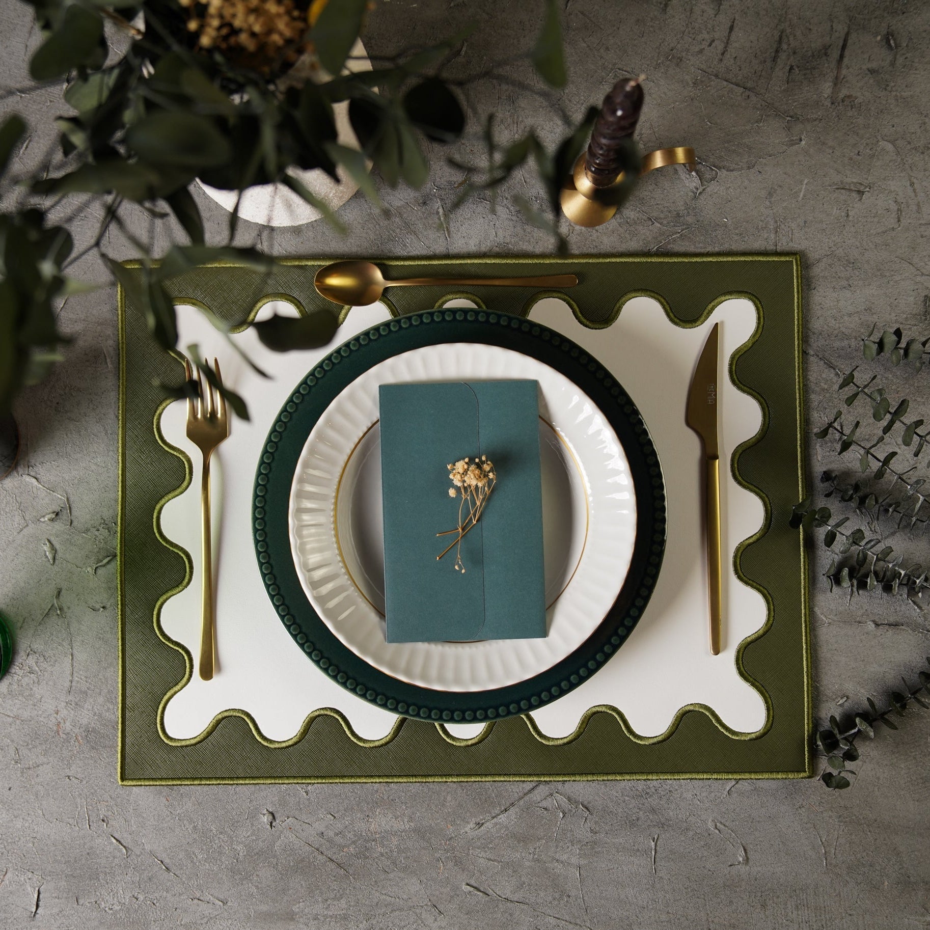 A white and dark green faux leather placemat with embroidered details, placed on a table with a plate and cutlery set on top.