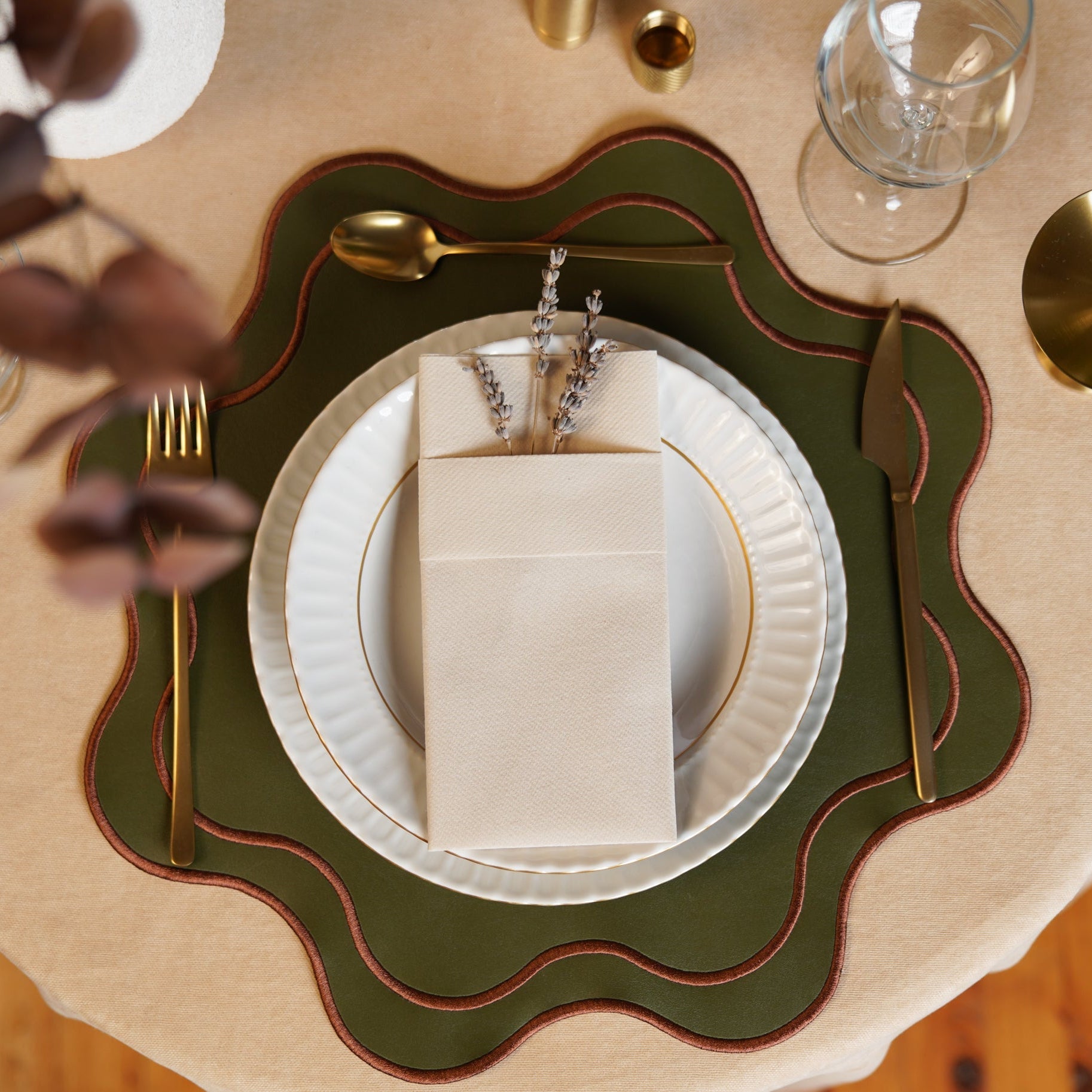 A round placemat with dark green and brown faux leather, placed under a white plate with a gold fork and spoon on a wooden table.
