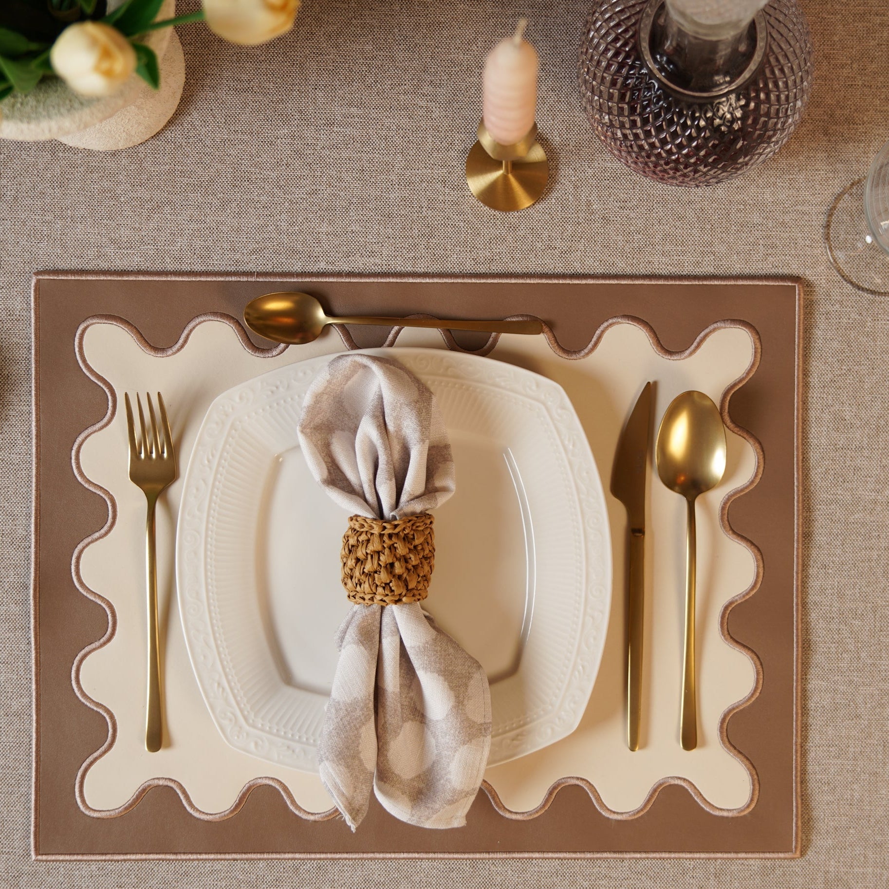 A brown faux leather placemat with embroidered details, placed on a dining table setting with cutlery and a napkin.
