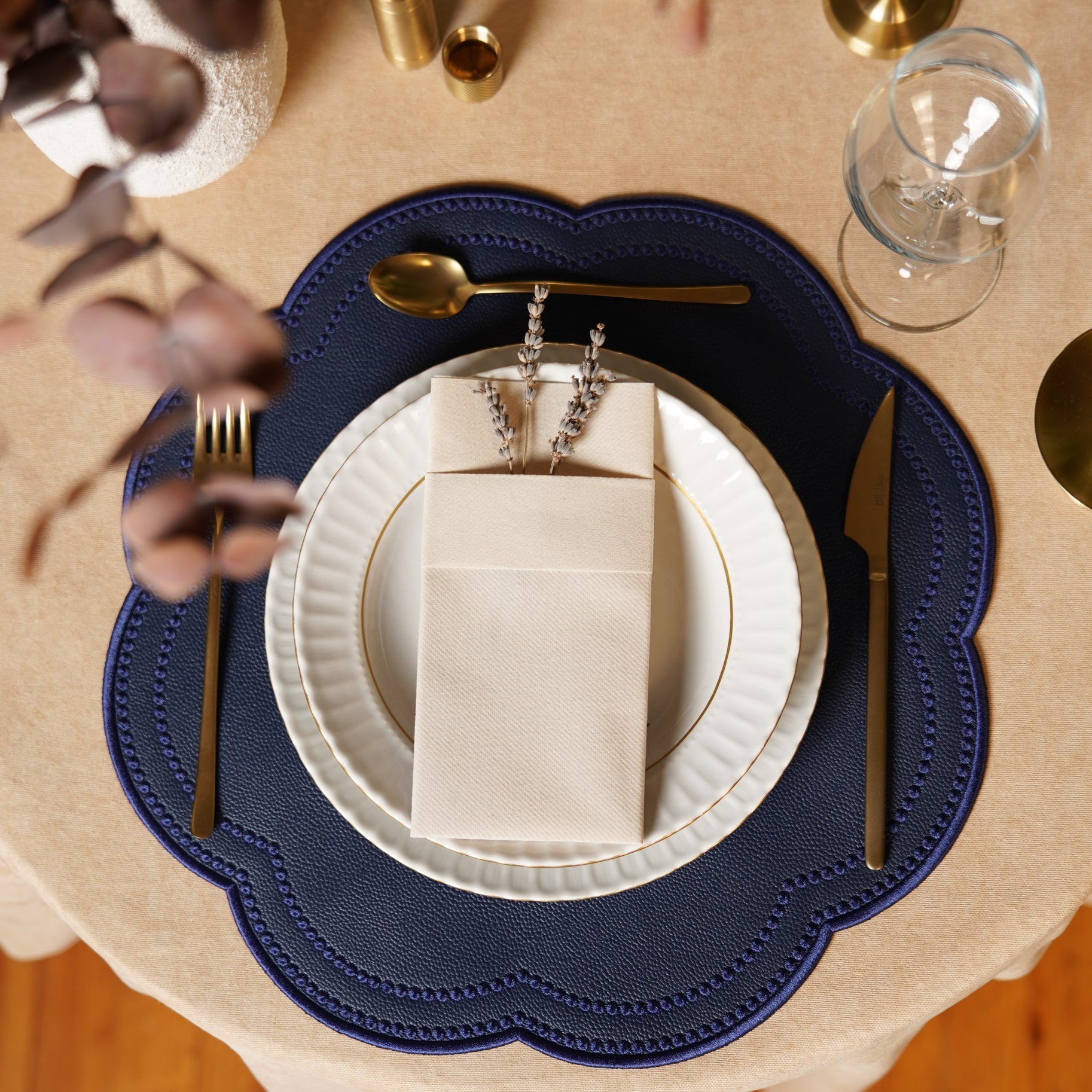 A navy blue faux leather placemat with a round edge design, placed on a dining table with a white plate, cutlery, and a glass on top of it.