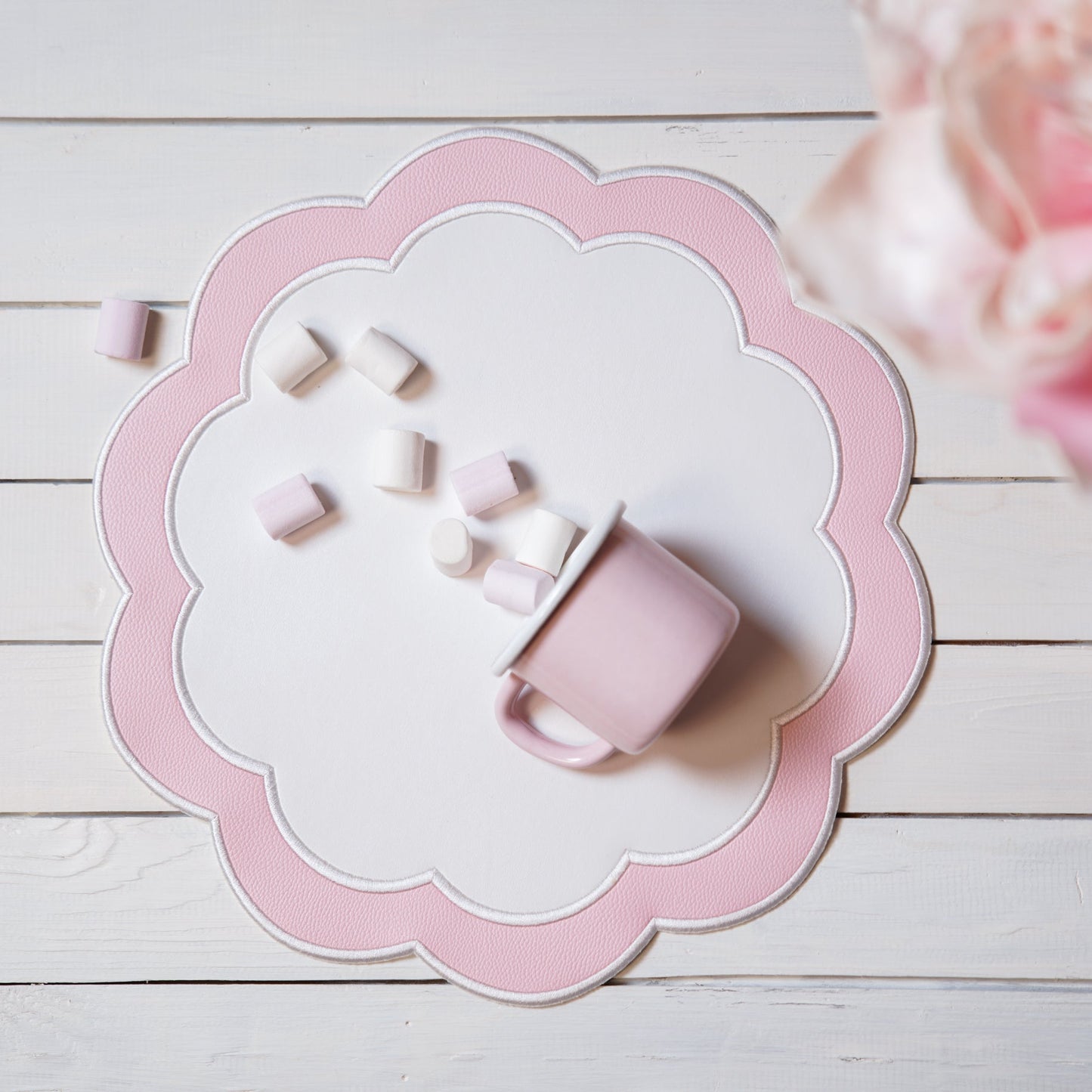 A round white faux leather placemat with pink embroidery, placed on a wooden surface with a pink flower in the background.