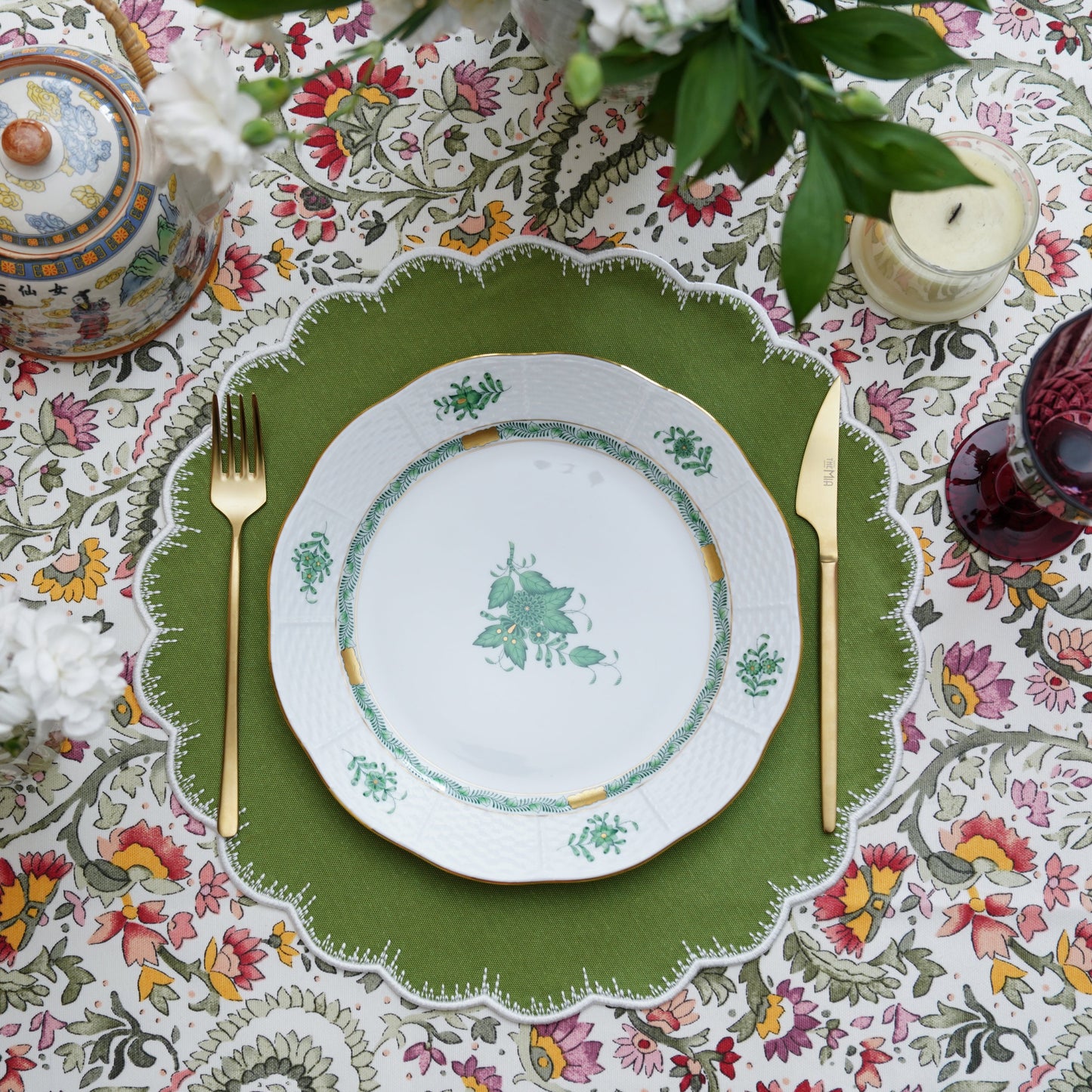 A set table with a green fabric placemat featuring a white trim, surrounded by a floral tablecloth, a white plate with green floral embroidery, and a teapot.