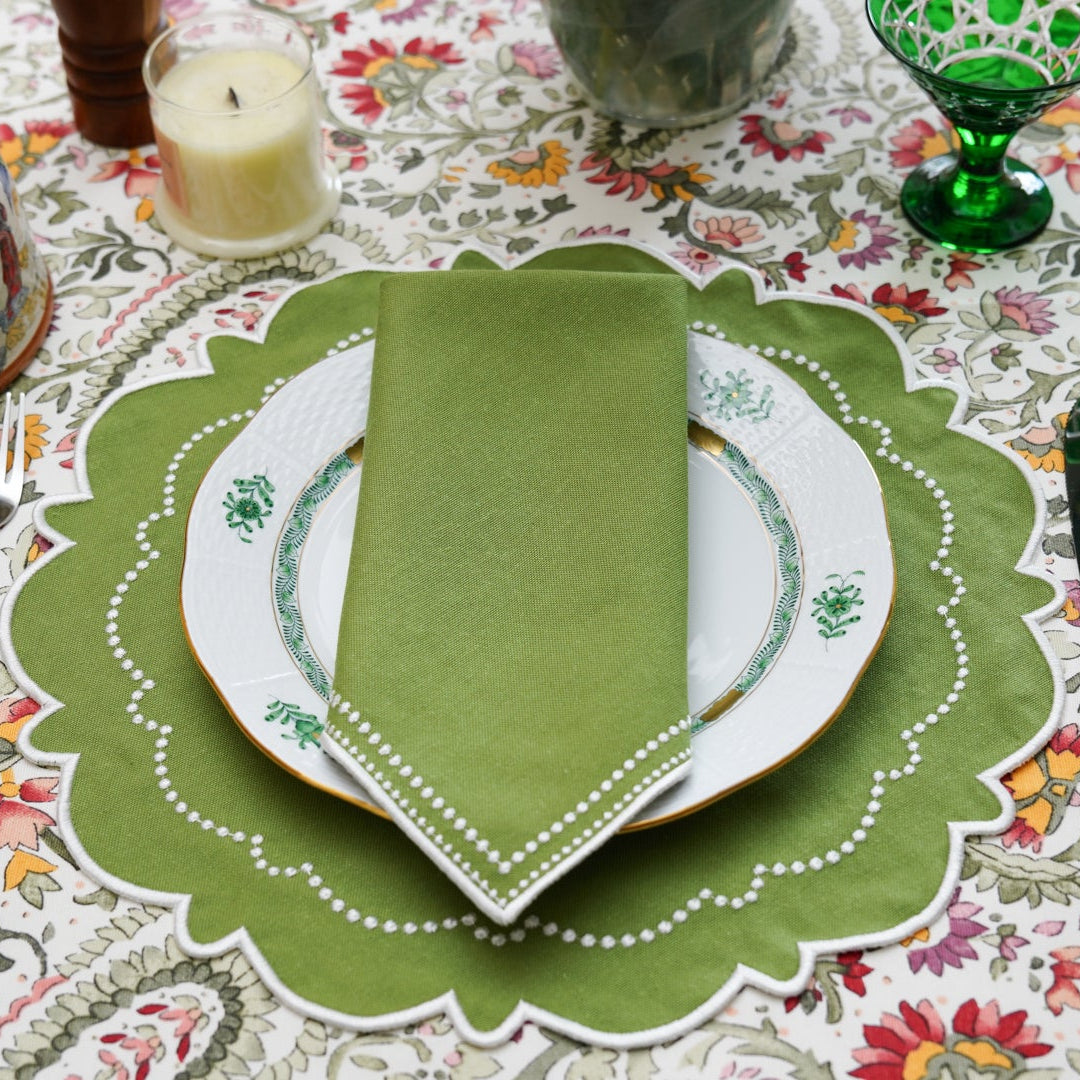 A green fabric placemat with white embroidery, displayed on a dining table with a napkin and cutlery.