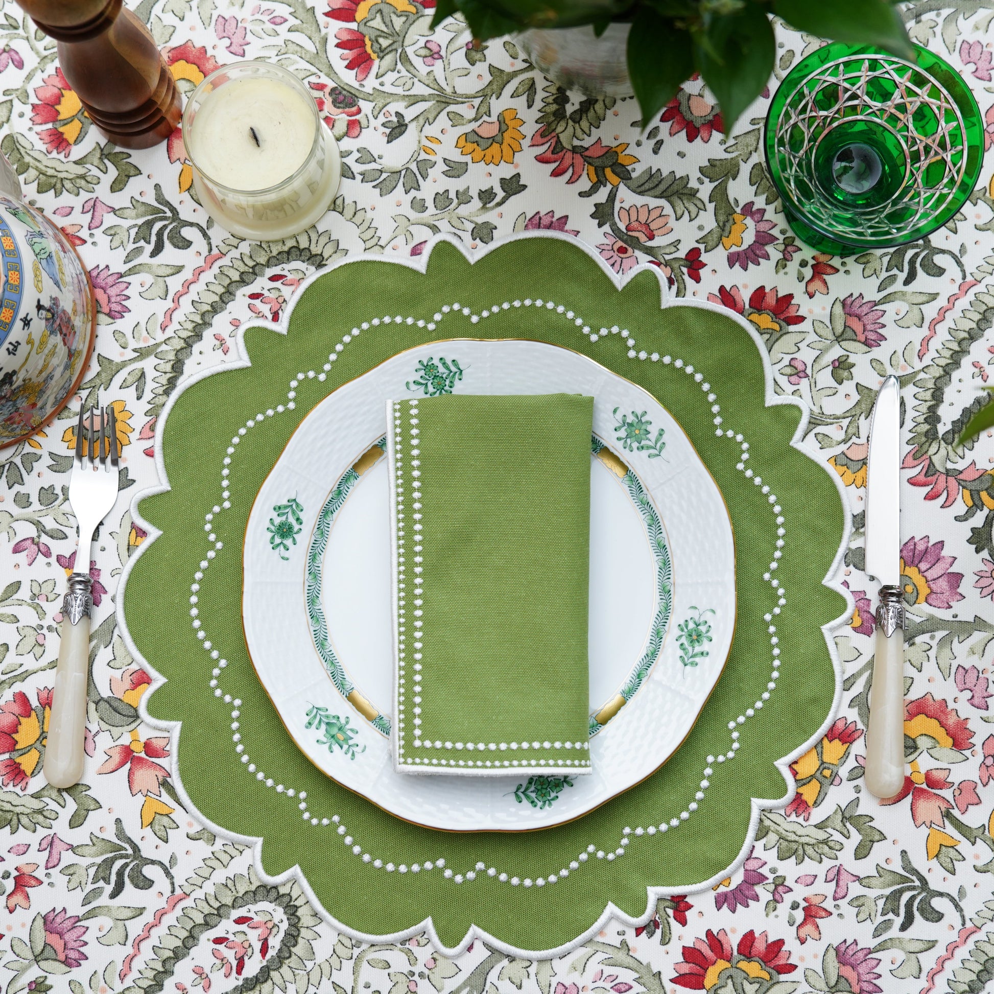 A set of green fabric placemats with white embroidery, displayed on a table with a floral tablecloth, plates, and cutlery.