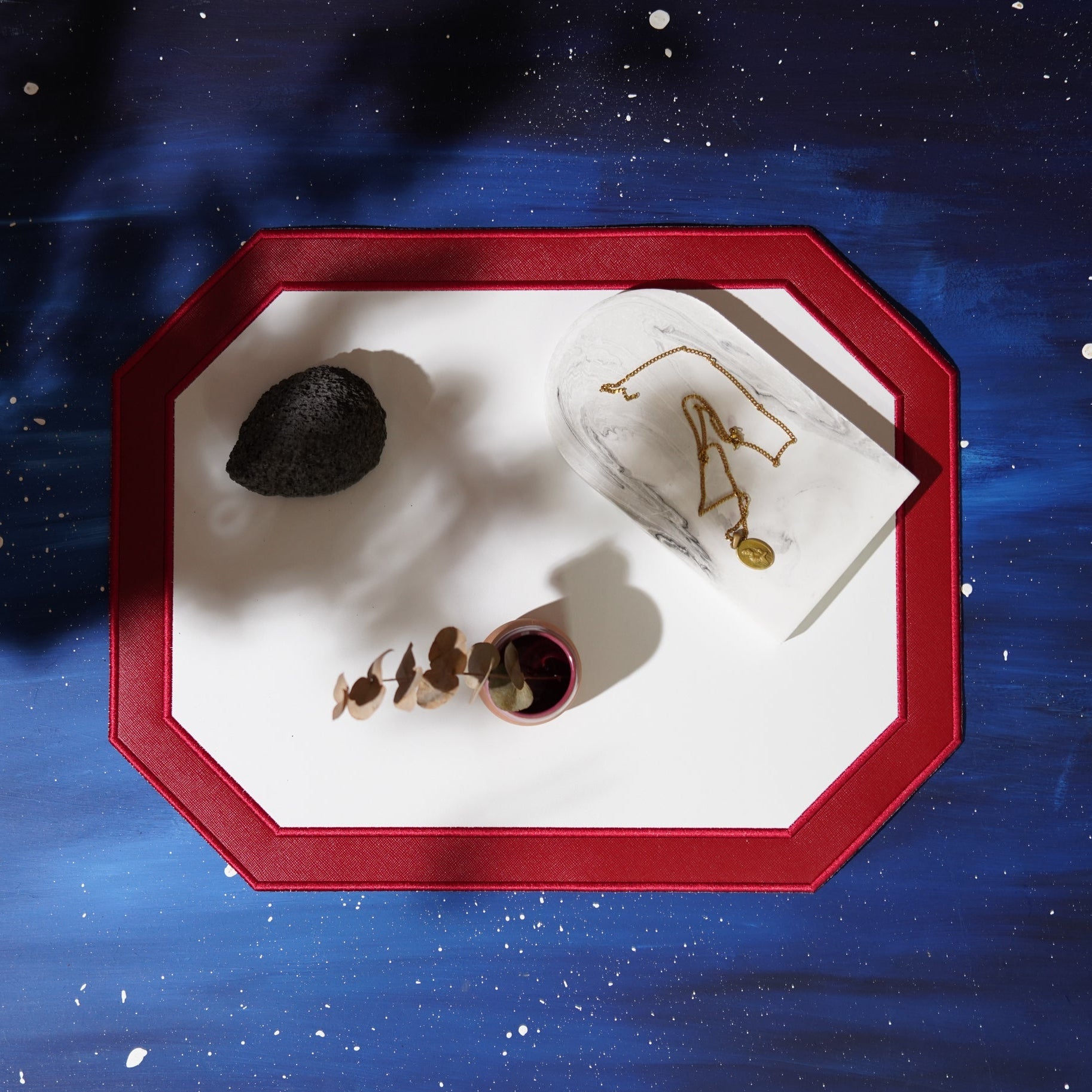 Decorative tray with a black stone, small plant, and gold necklace against a starry night sky background.