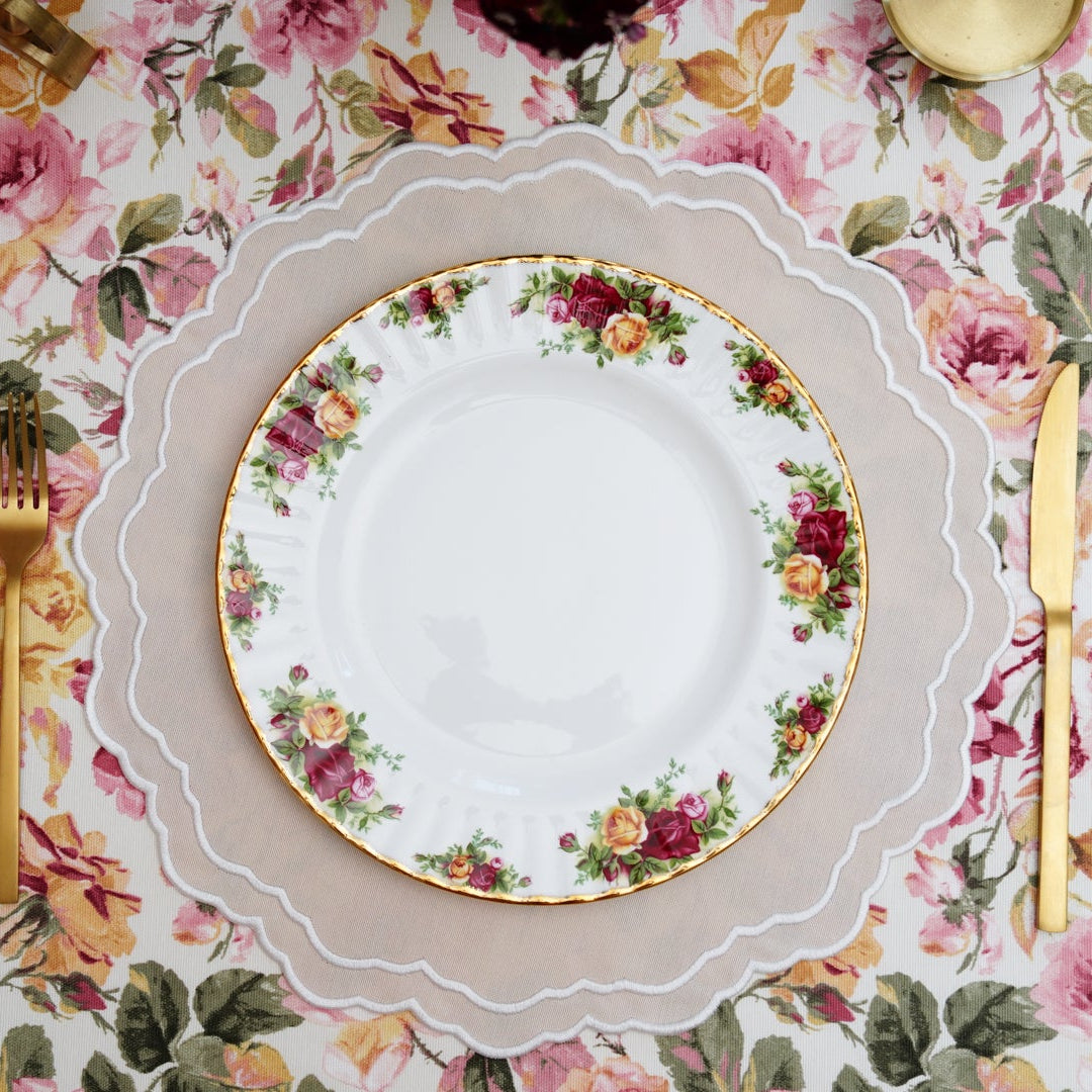 A round beige fabric placemat with white trim, laid out on a table with a floral pattern. A plate and cutlery are placed on top of the placemat.