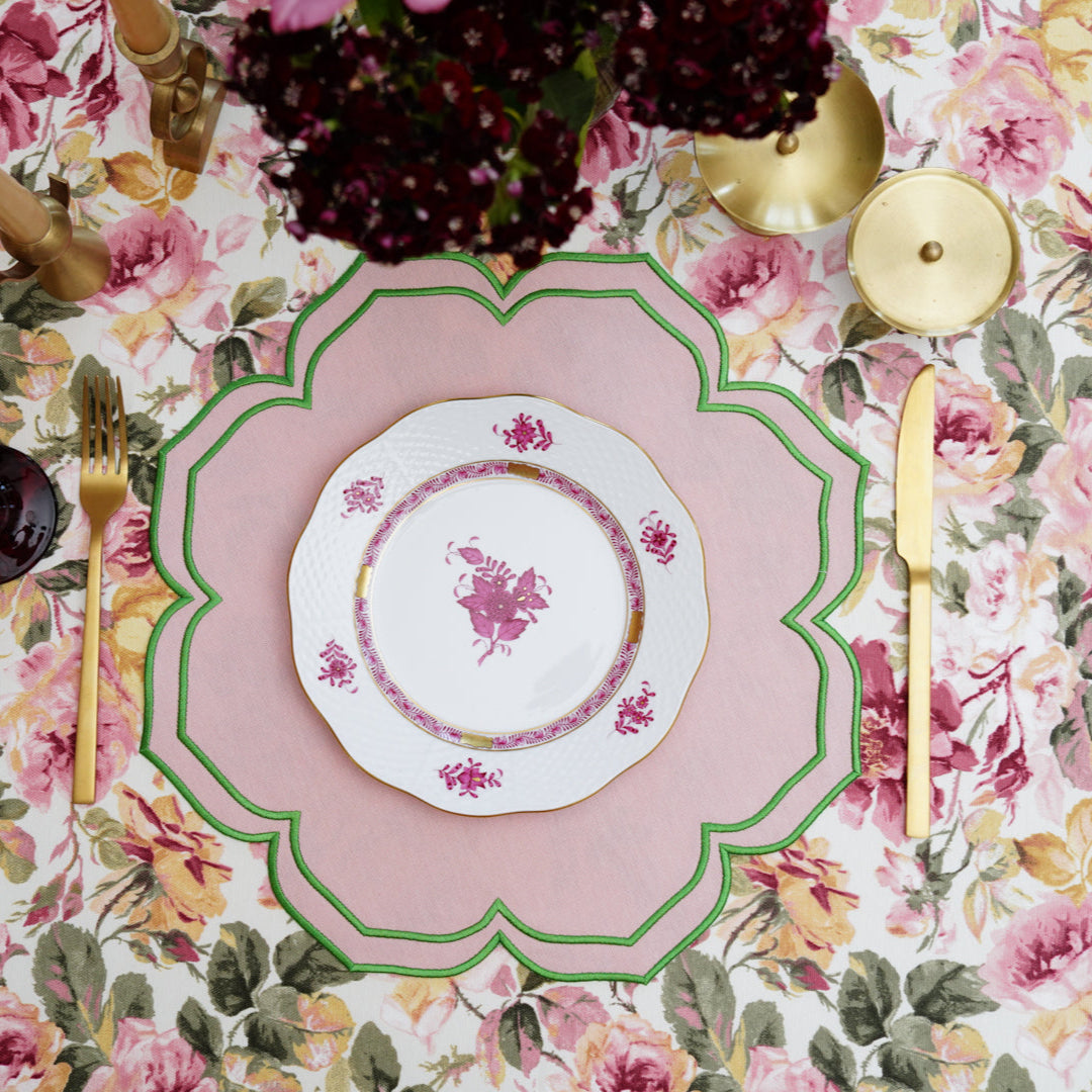 A round pink and green fabric placemat with floral embroidery, displayed on a table with cutlery and a floral tablecloth.