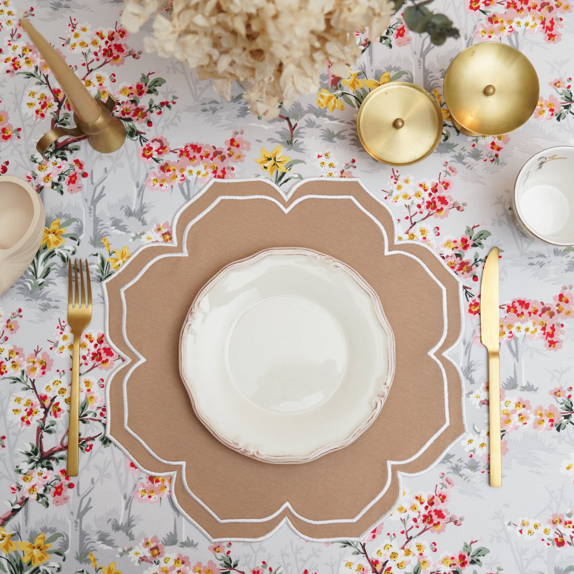 A beige fabric placemat with white trim, displayed on a table with a floral tablecloth, cutlery, and decorative items.