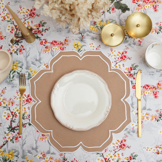 A beige fabric placemat with white trim, displayed on a table with a floral tablecloth, cutlery, and decorative items.