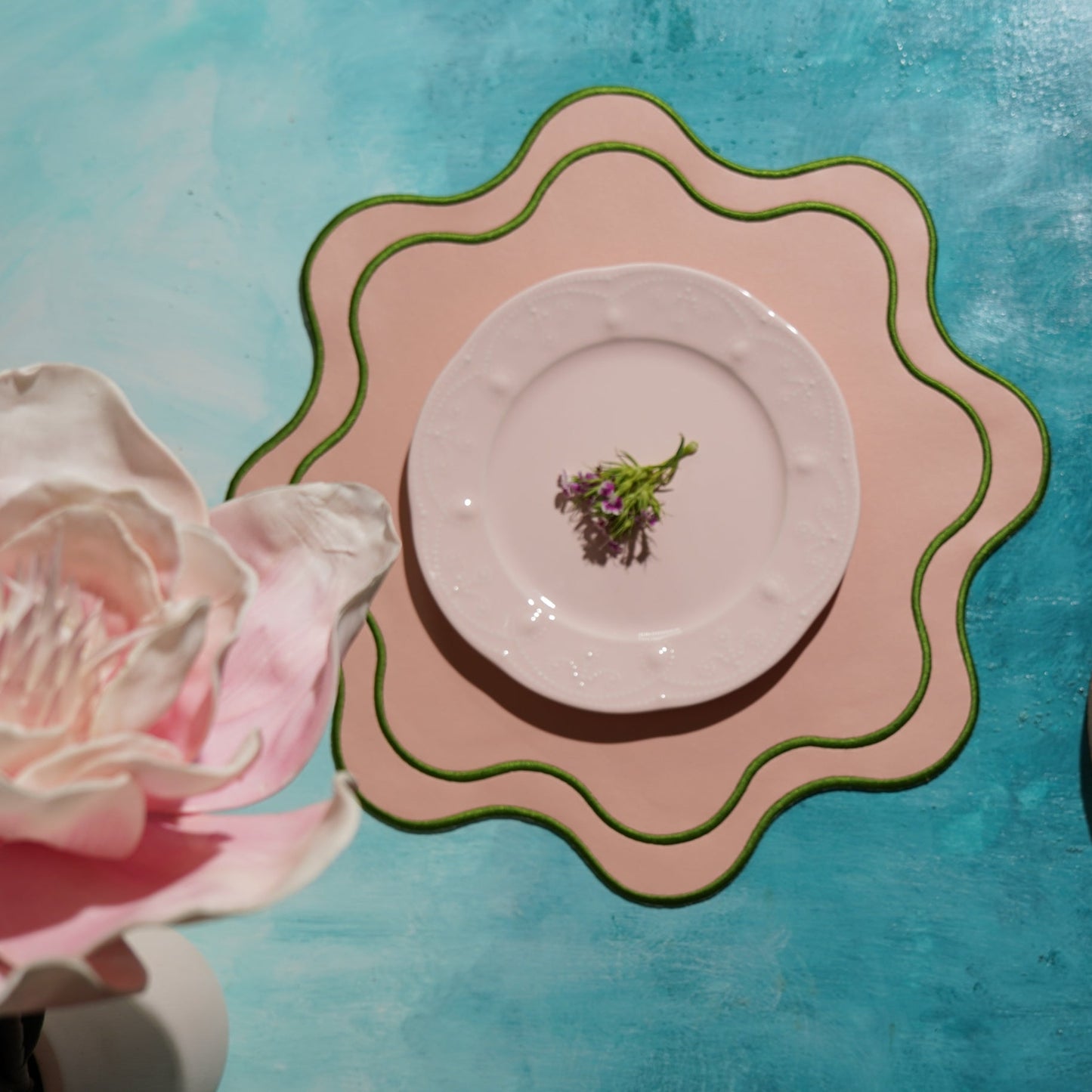A pink faux leather placemat with a decorative edge, presented on a table with a plate and cutlery setup, surrounded by a blue backdrop and a pink rose to the side.
