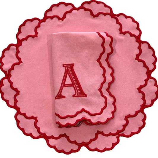 A pink fabric placemat with a red embroidered letter 'A' in the center, designed with a scalloped border.