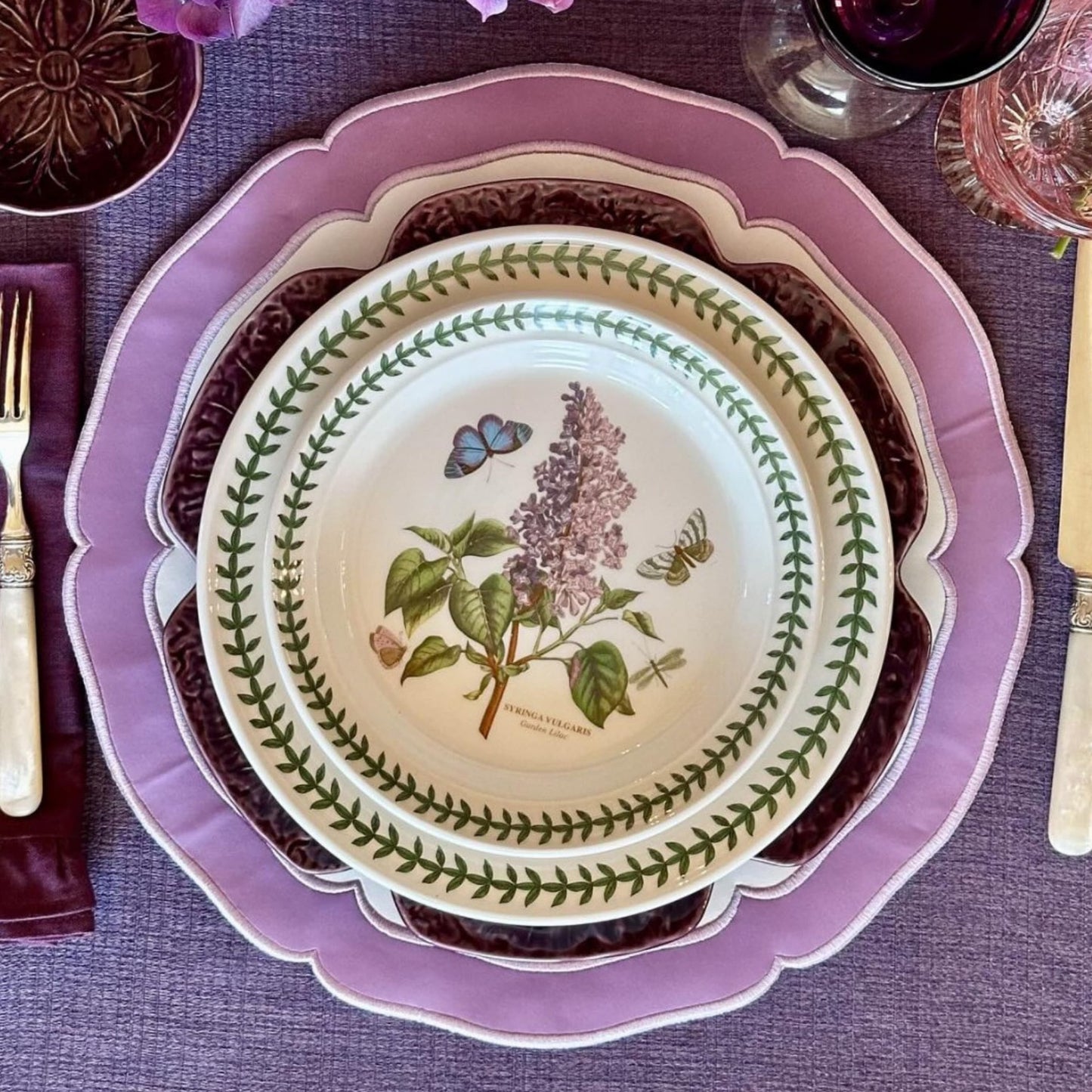 A round faux leather placemat in white and purple with floral patterns, displayed on a dining table setting with cutlery and a purple napkin.