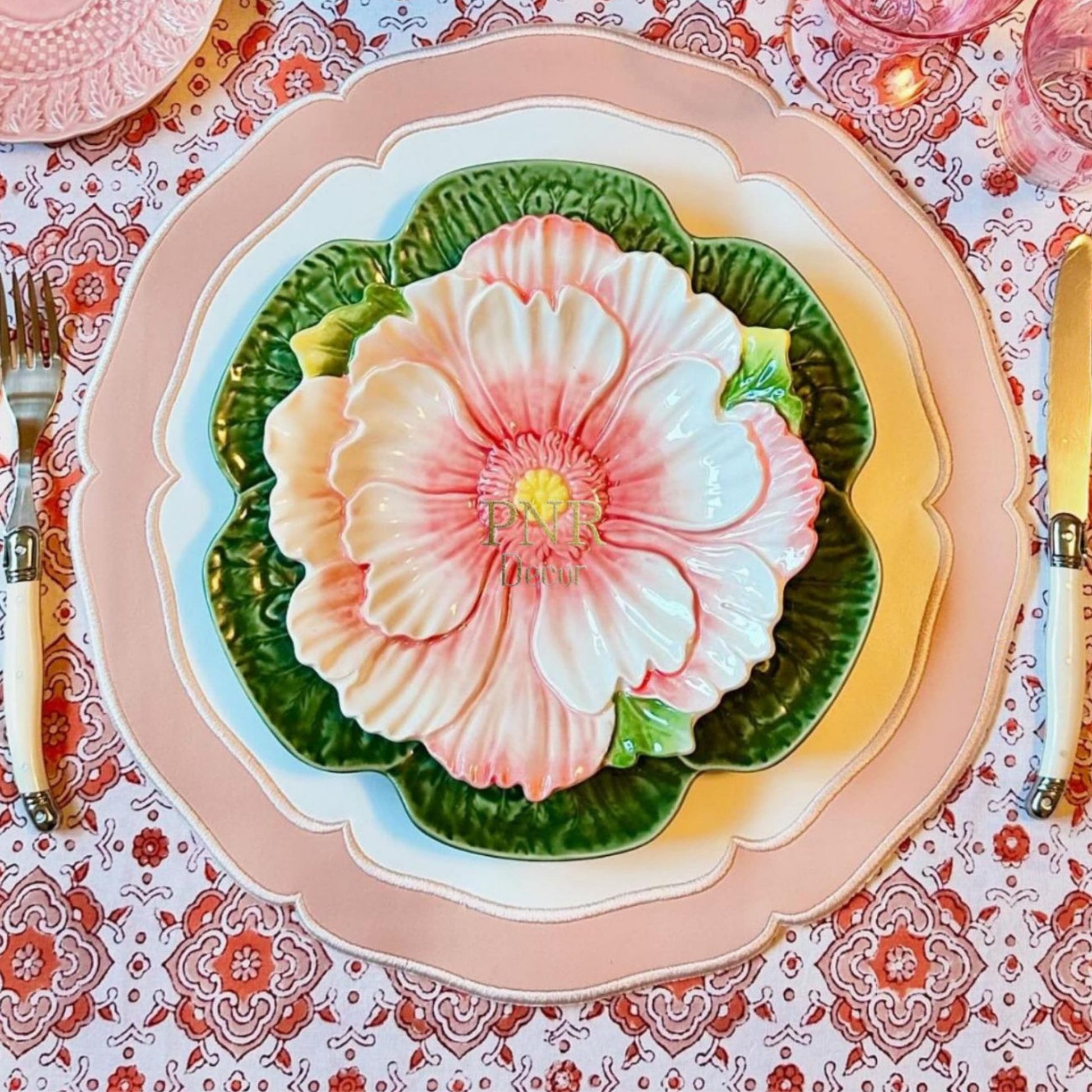 A round placemat with a floral design in white and pink colors, displayed on a table with cutlery and a patterned tablecloth.