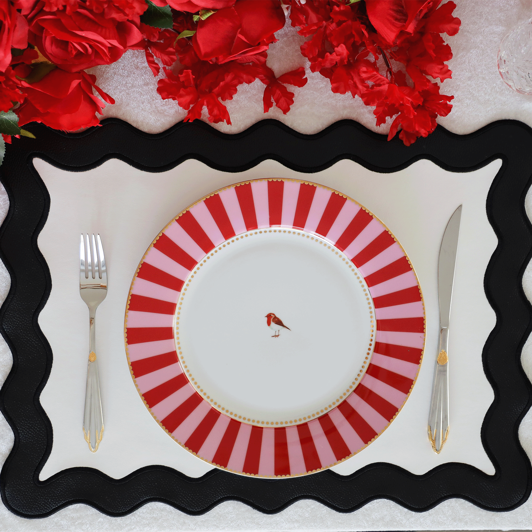 A black and white faux leather placemat with a decorative edge, presented on a table with a red and white striped plate on top and red flowers in the background.