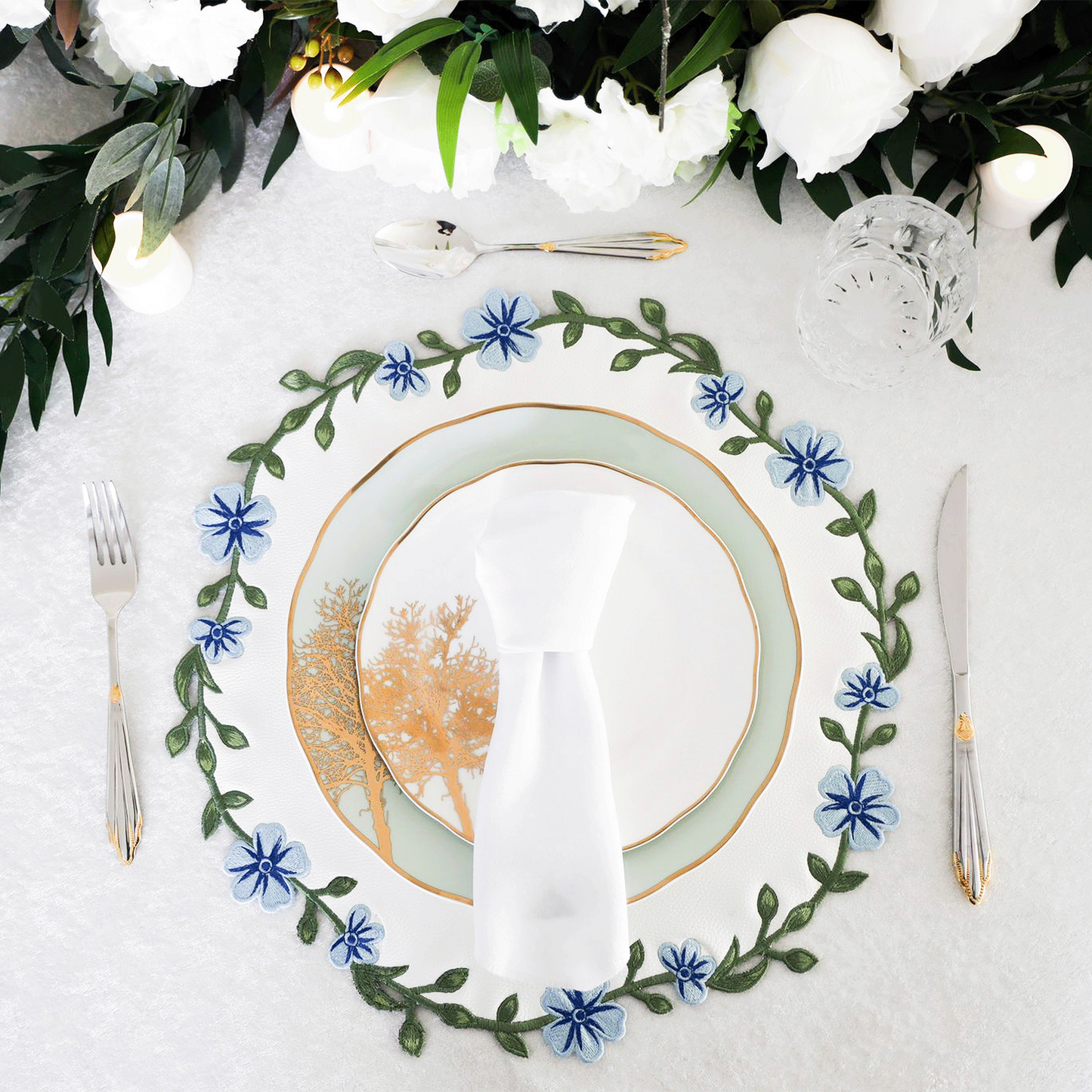 A round faux leather placemat with white and blue floral embroidery, displayed on a table setting with cutlery and a plate.