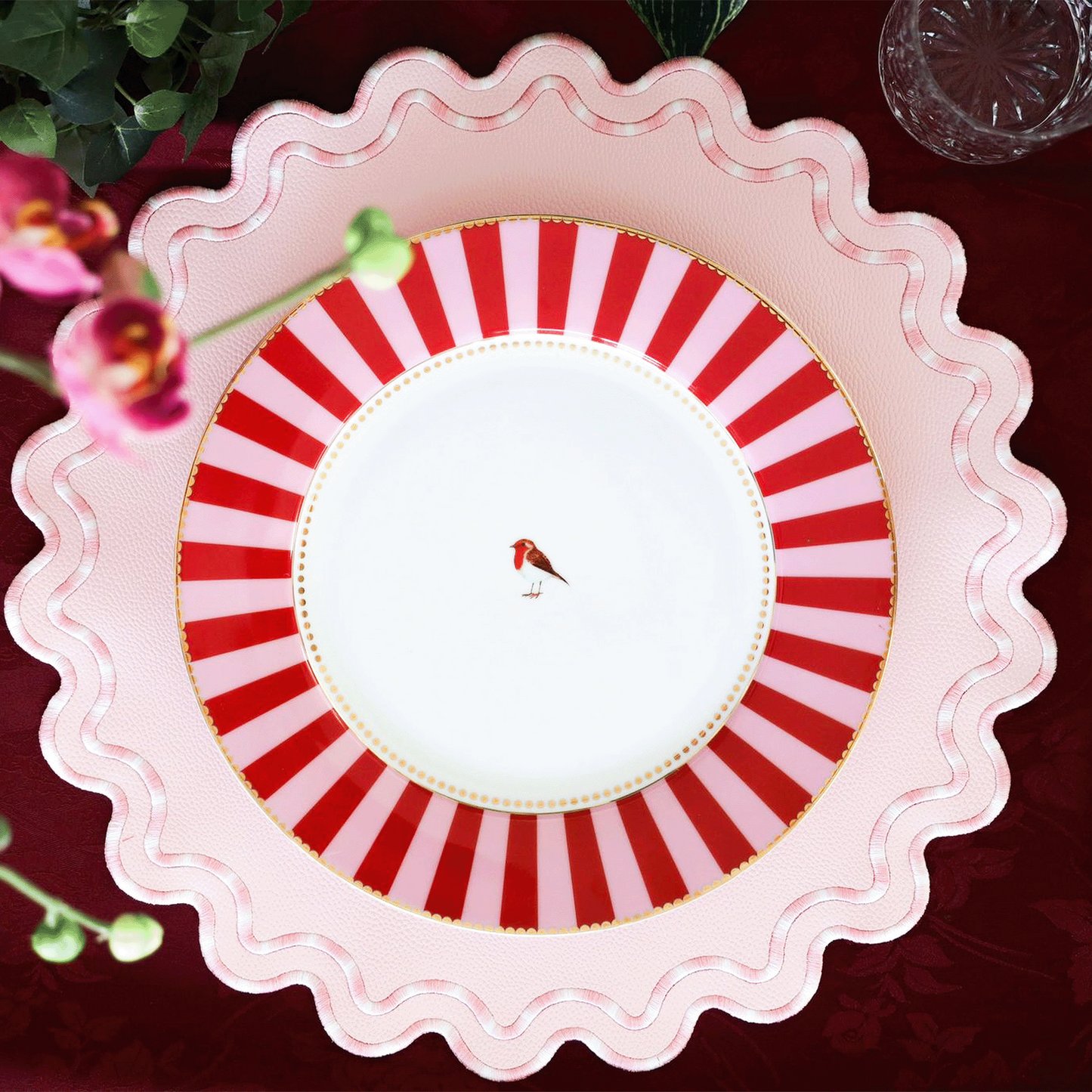 A round, pink faux leather placemat with a striped pattern, presented on a table setting with cutlery and a floral decoration.