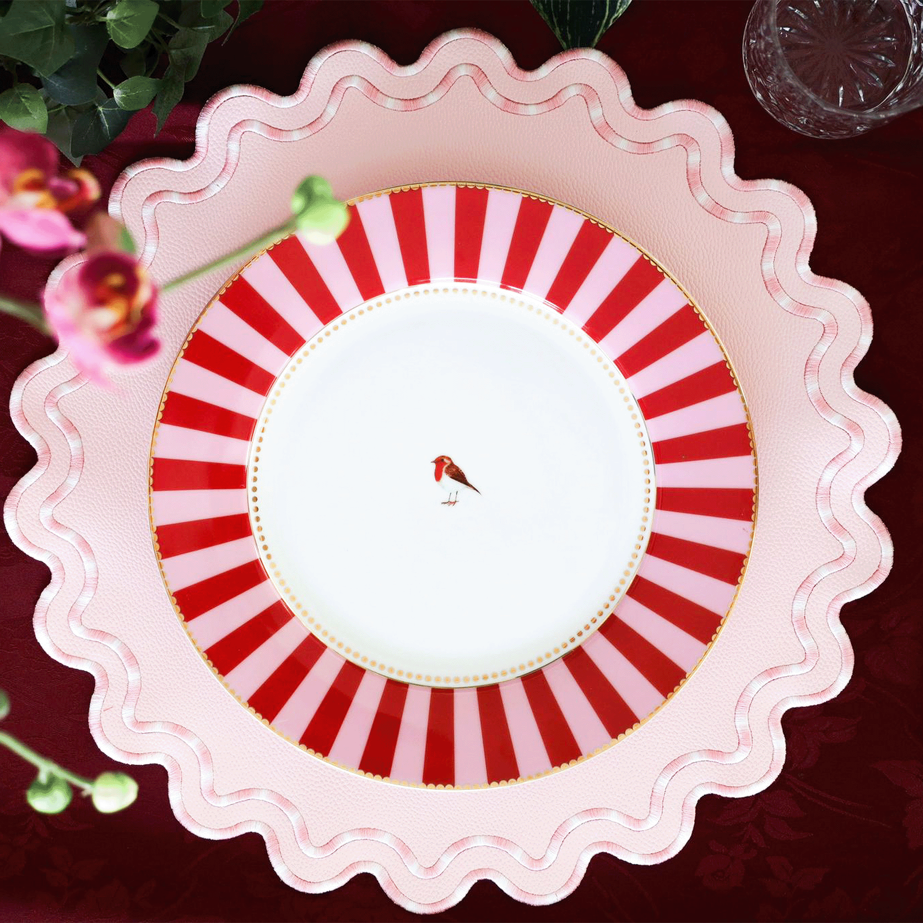A round, pink faux leather placemat with a striped pattern, presented on a table setting with cutlery and a floral decoration.