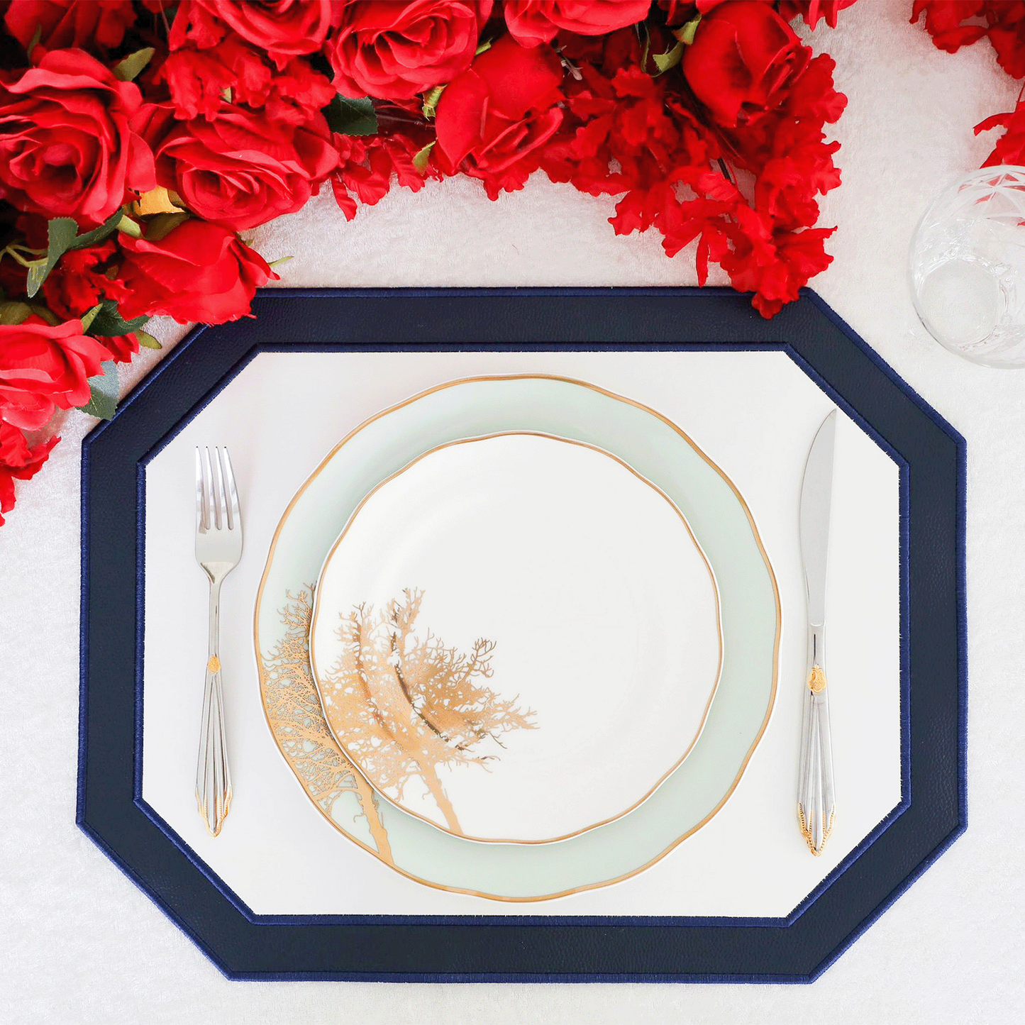 A navy blue faux leather placemat with white embroidered detailing, presented on a table with a plate and cutlery, surrounded by red roses.