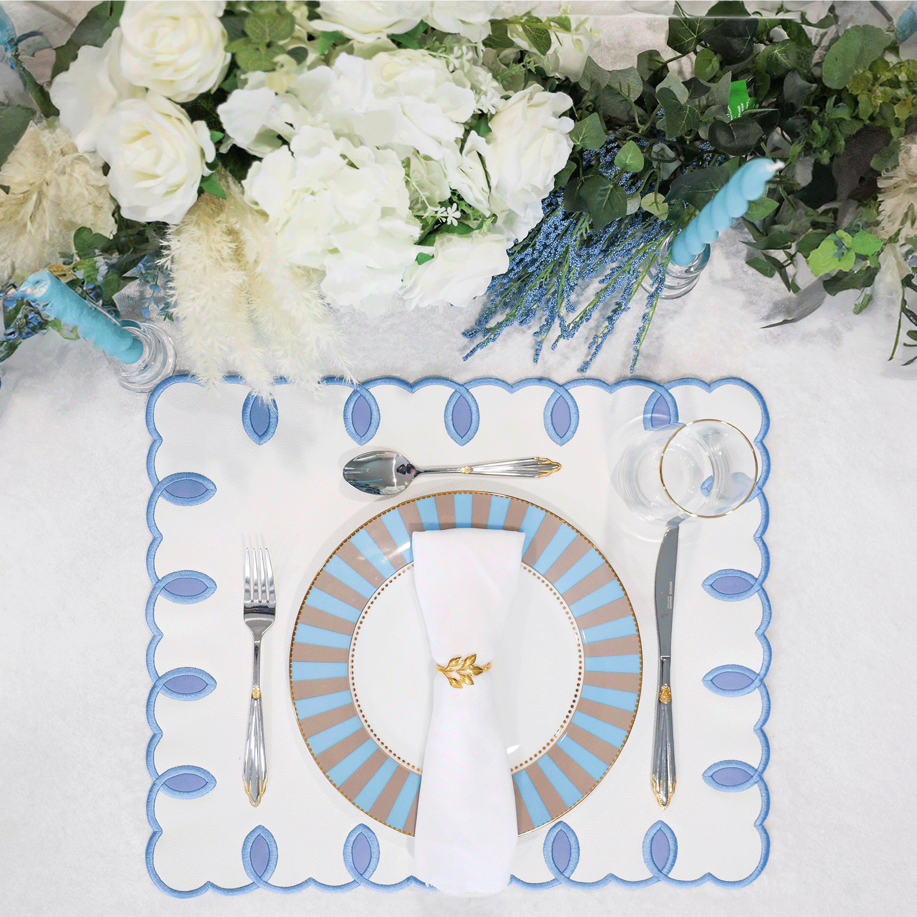 A set table with a white and blue embroidered faux leather placemat, a white plate, and silverware. The table is set against a floral backdrop.