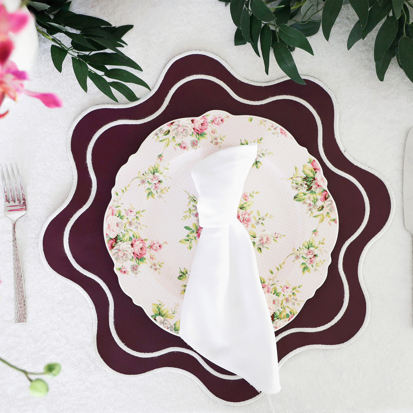 A burgundy faux leather placemat with scalloped edges, presented on a table setting with a floral plate, silverware, and pink orchids.