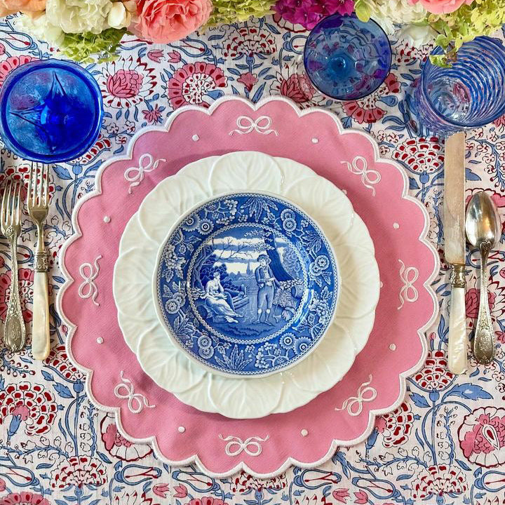Decorative table setting with pink and blue plates, flowers, and cutlery on a patterned tablecloth.