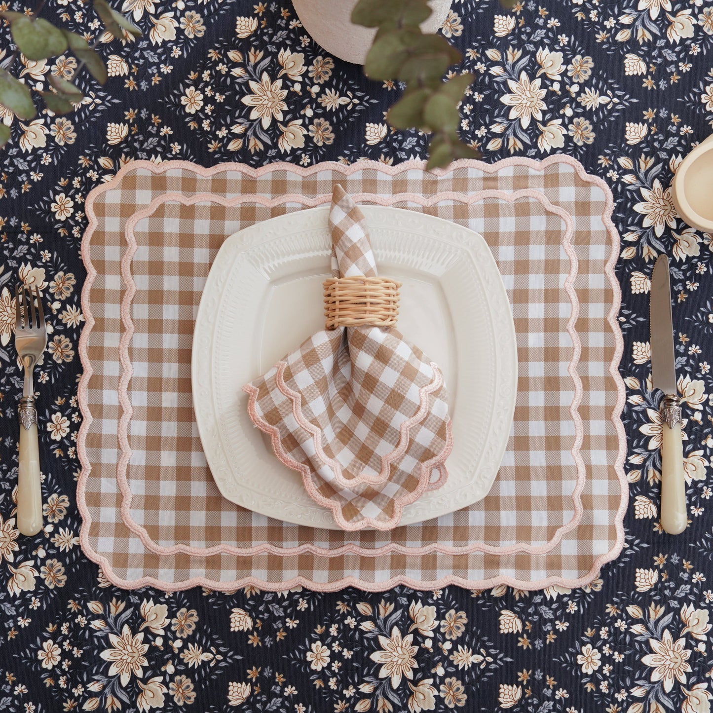 A beige checkered fabric placemat with pink trim, displayed on a floral patterned tablecloth. A napkin is also visible, tied with a matching checkered fabric.