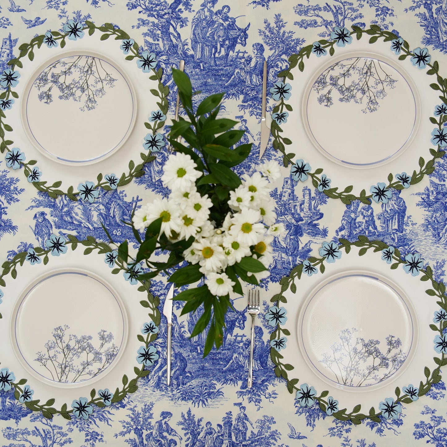 Table setting with white plates, silverware, and a floral centerpiece on a blue and white toile tablecloth.