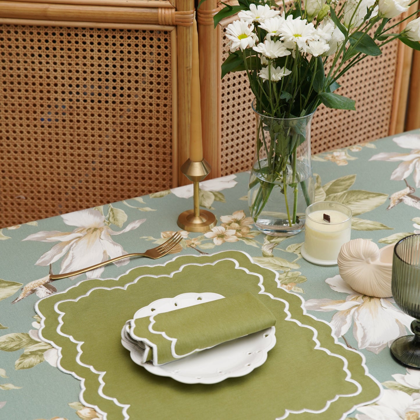 Set table with floral tablecloth, green placemat, and white flowers in a vase.