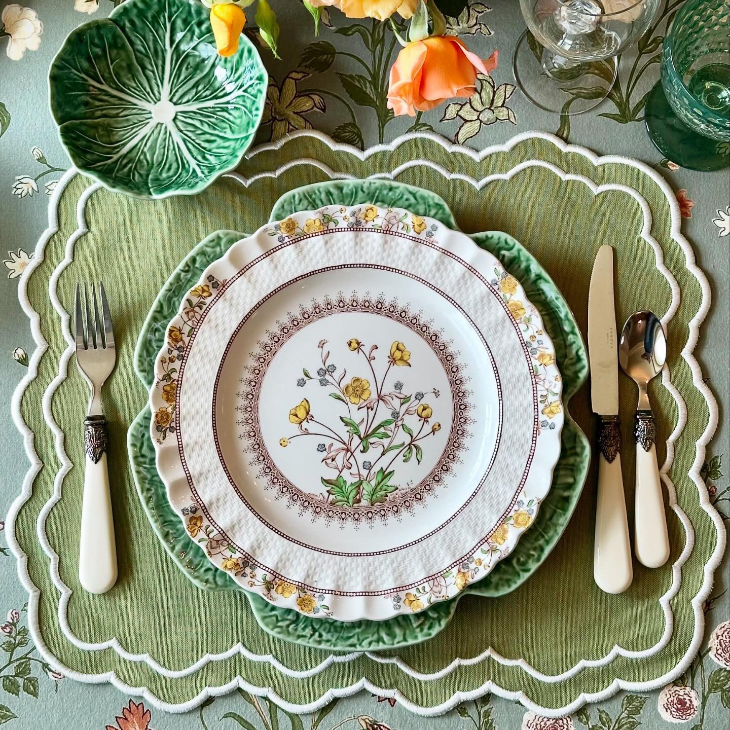 Decorative table setting with floral plates, cutlery, and flowers on a patterned tablecloth.