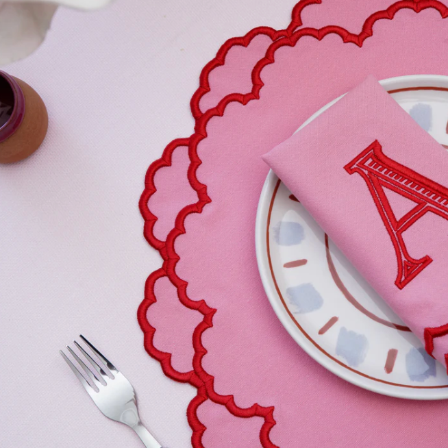 Pink placemat with scalloped edge, white plate, and pink napkin with letter 'A' on a white table.