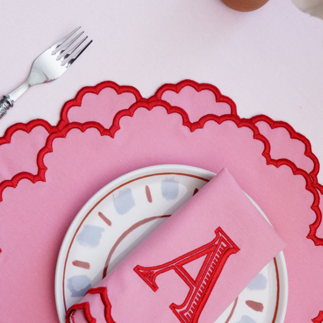 Pink placemat with scalloped edge, white plate with blue pattern, and pink napkin with letter 'A' on a white surface.