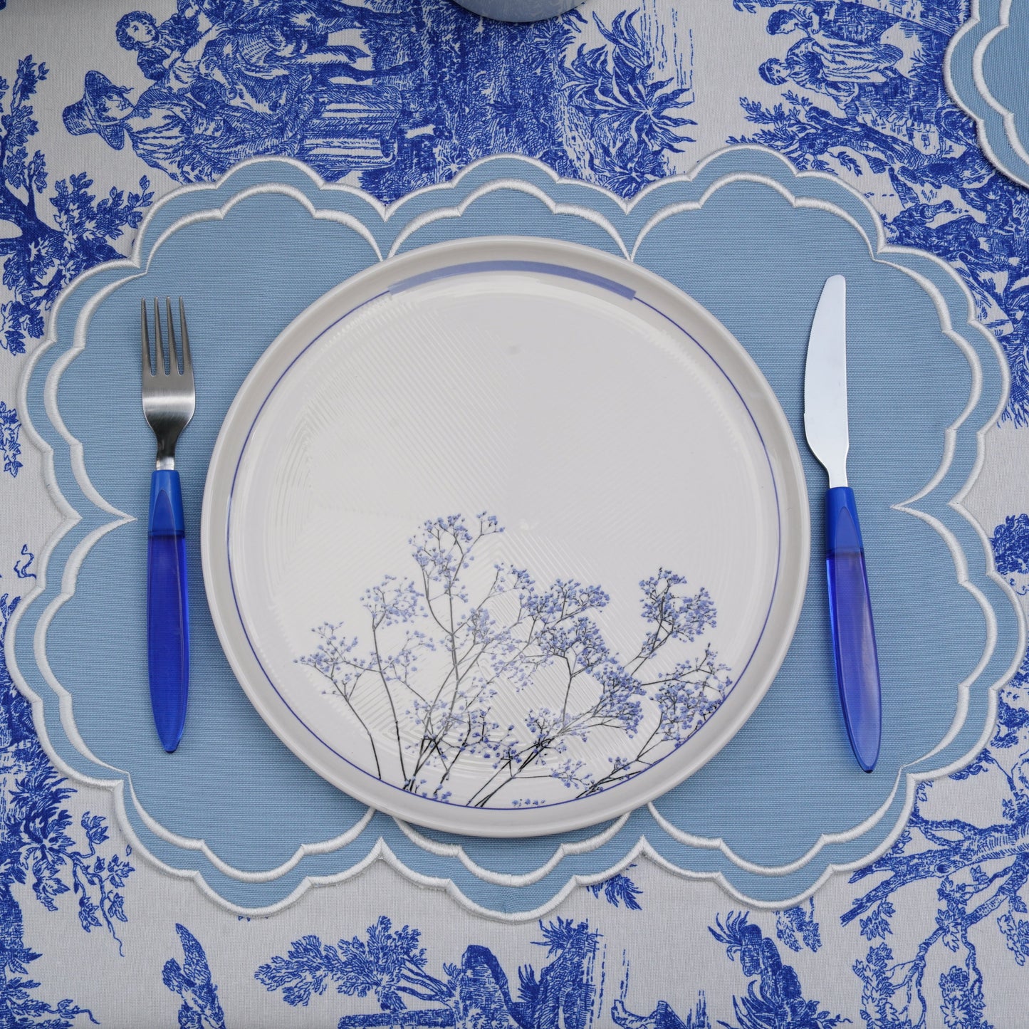 A blue and white floral patterned placemat with a plate and cutlery set on top of it, displayed on a table.