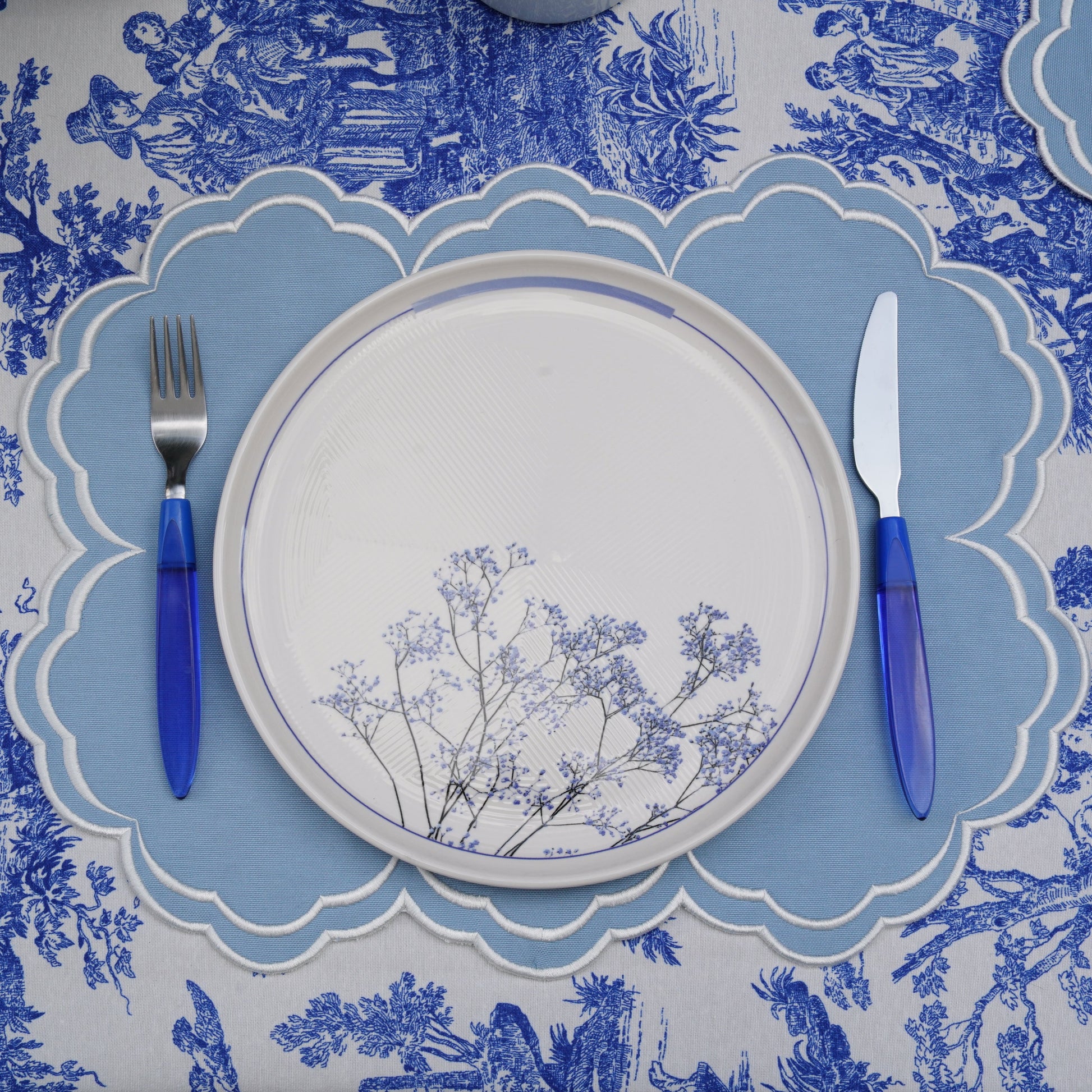 A blue and white floral patterned placemat with a plate and cutlery set on top of it, displayed on a table.